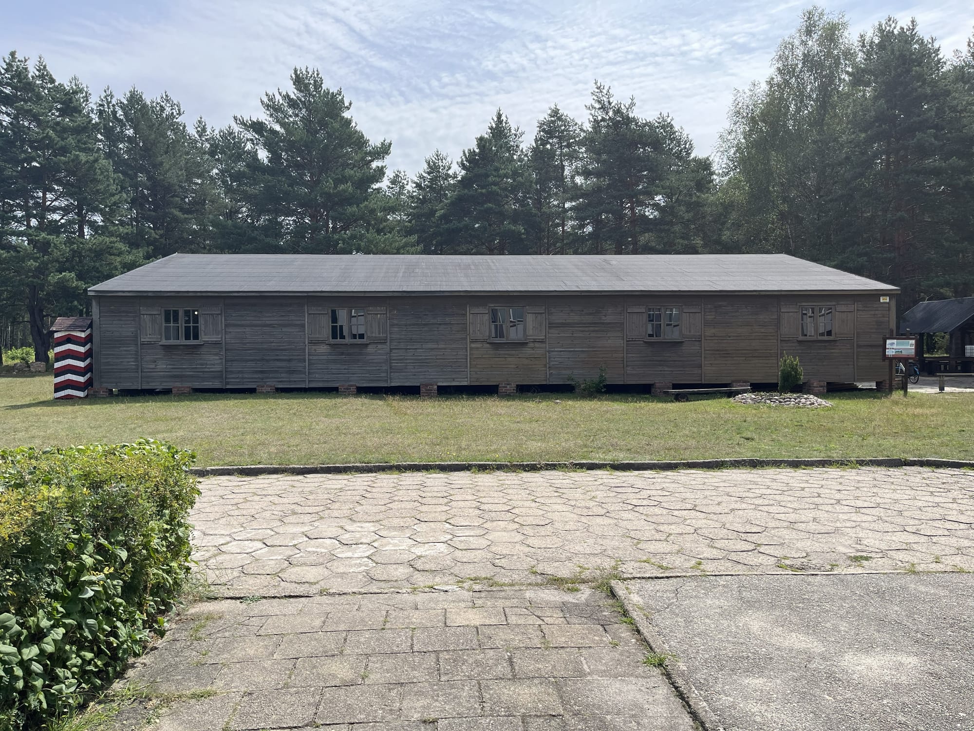 Reconstructed wooden barrack at the former Stalag Luft III prisoner-of-war camp, standing in a clearing surrounded by pine trees beneath a bright sky.