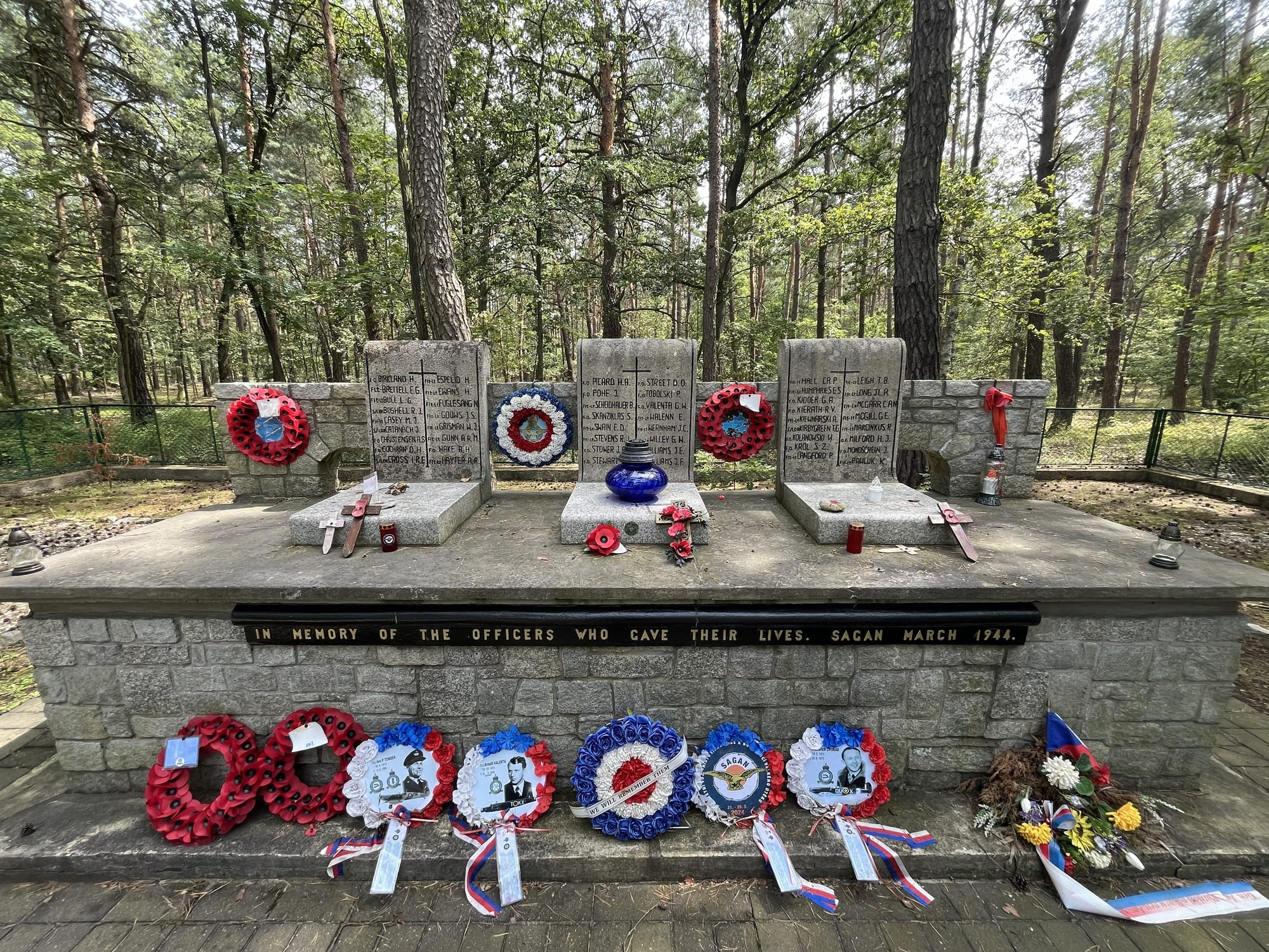 Memorial at the former Stalag Luft III camp featuring engraved stone plaques, remembrance wreaths, candles and tributes, set within a pine forest clearing.