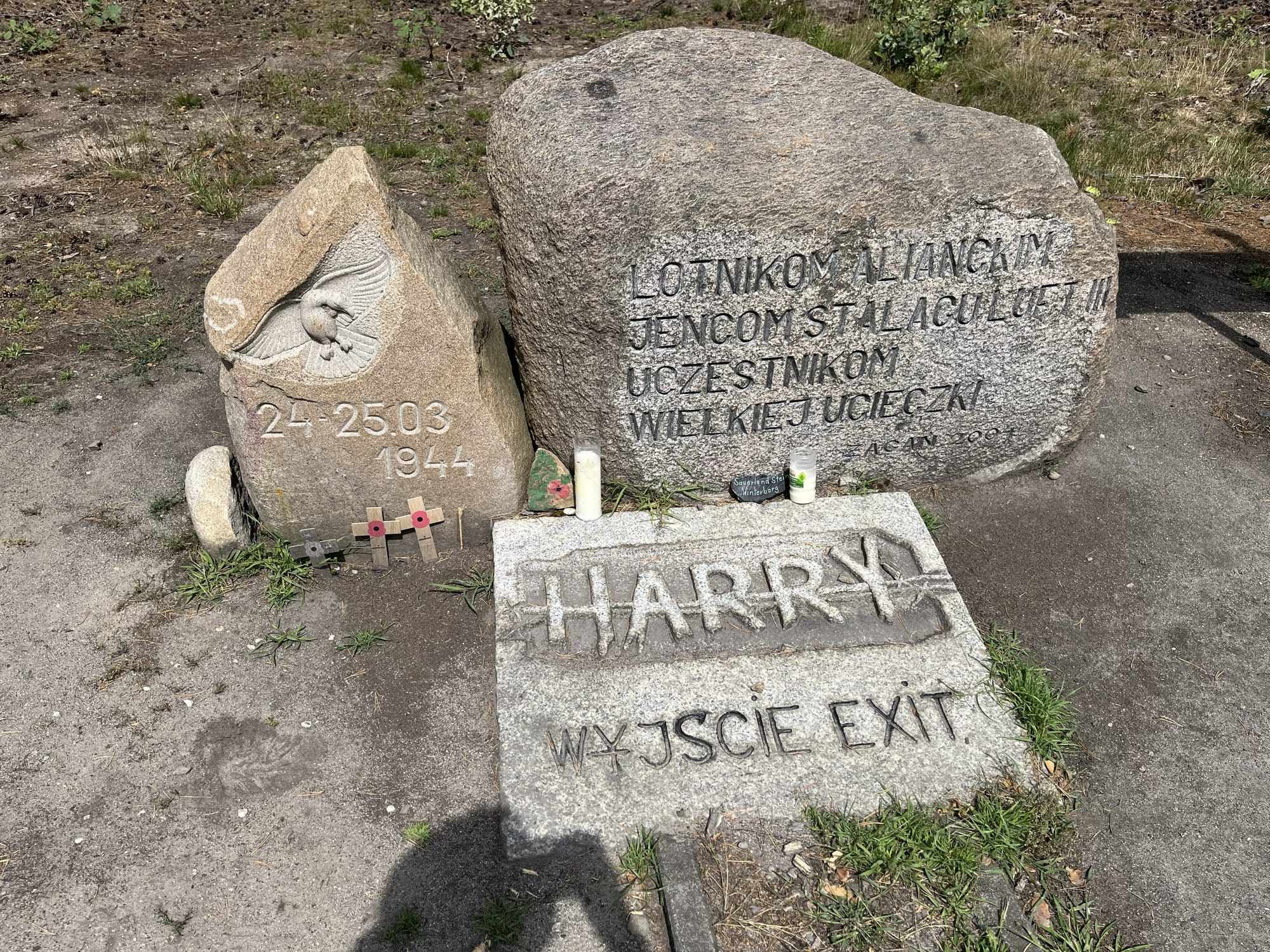 Memorial stones marking the exit of Tunnel Harry at the former Stalag Luft III prisoner-of-war camp, featuring engraved inscriptions, remembrance candles and small commemorative crosses.