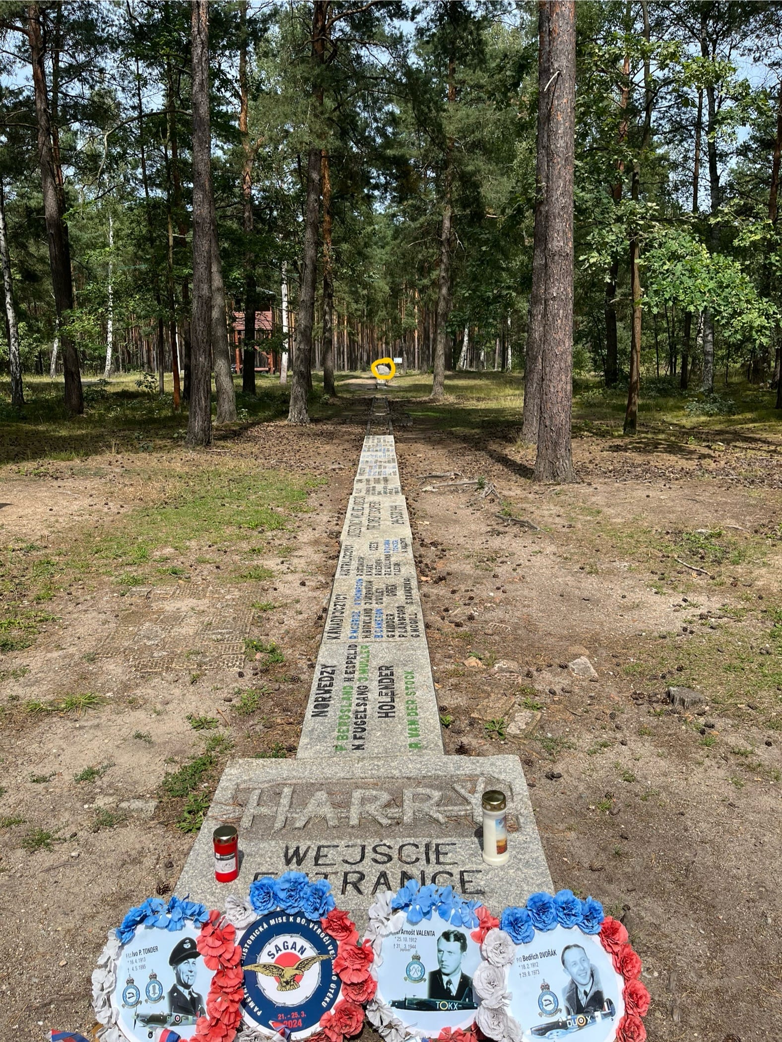 Memorial marking the entrance to Tunnel Harry at the former Stalag Luft III camp, with commemorative wreaths and candles beside a long stone marker extending through a pine forest towards the tunnel site.