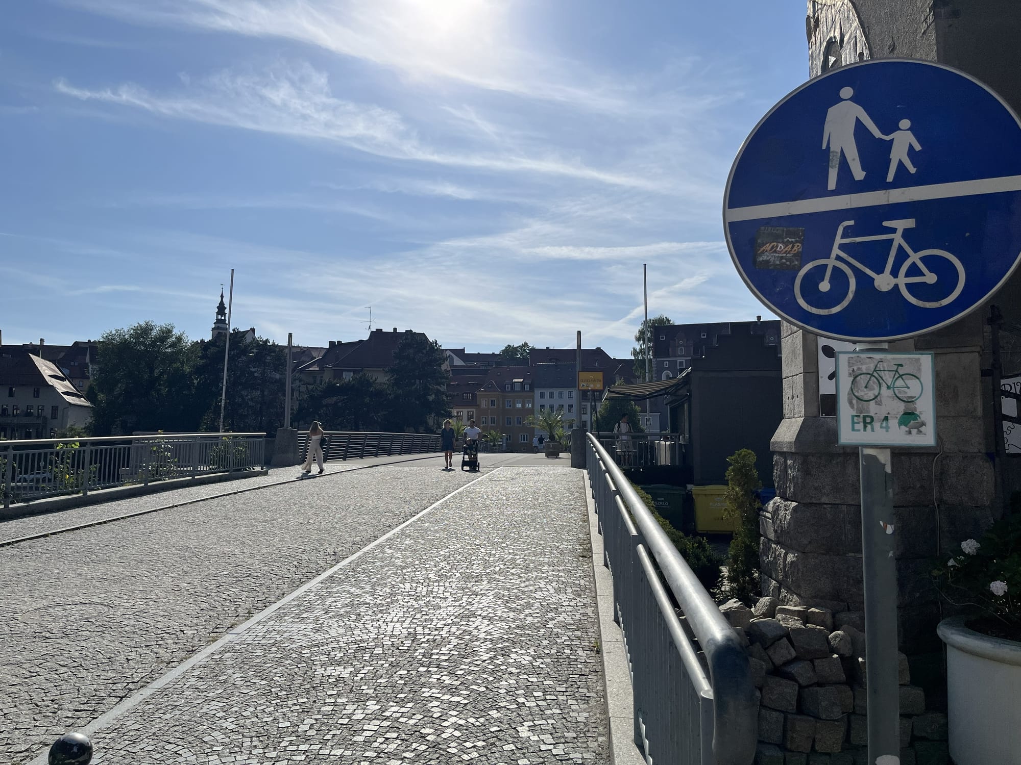 The Old Town Bridge linking Zgorzelec, Poland, and Görlitz, Germany, with a blue shared pedestrian and cycle path sign in the foreground and historic town buildings visible beyond under a bright summer sky.