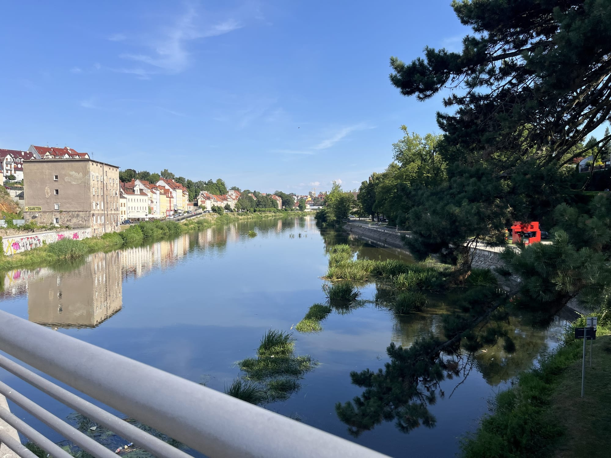 View from the Old Town Bridge over the River Neisse between Zgorzelec and Görlitz, showing historic riverside buildings reflected in the calm water beneath a clear blue sky.