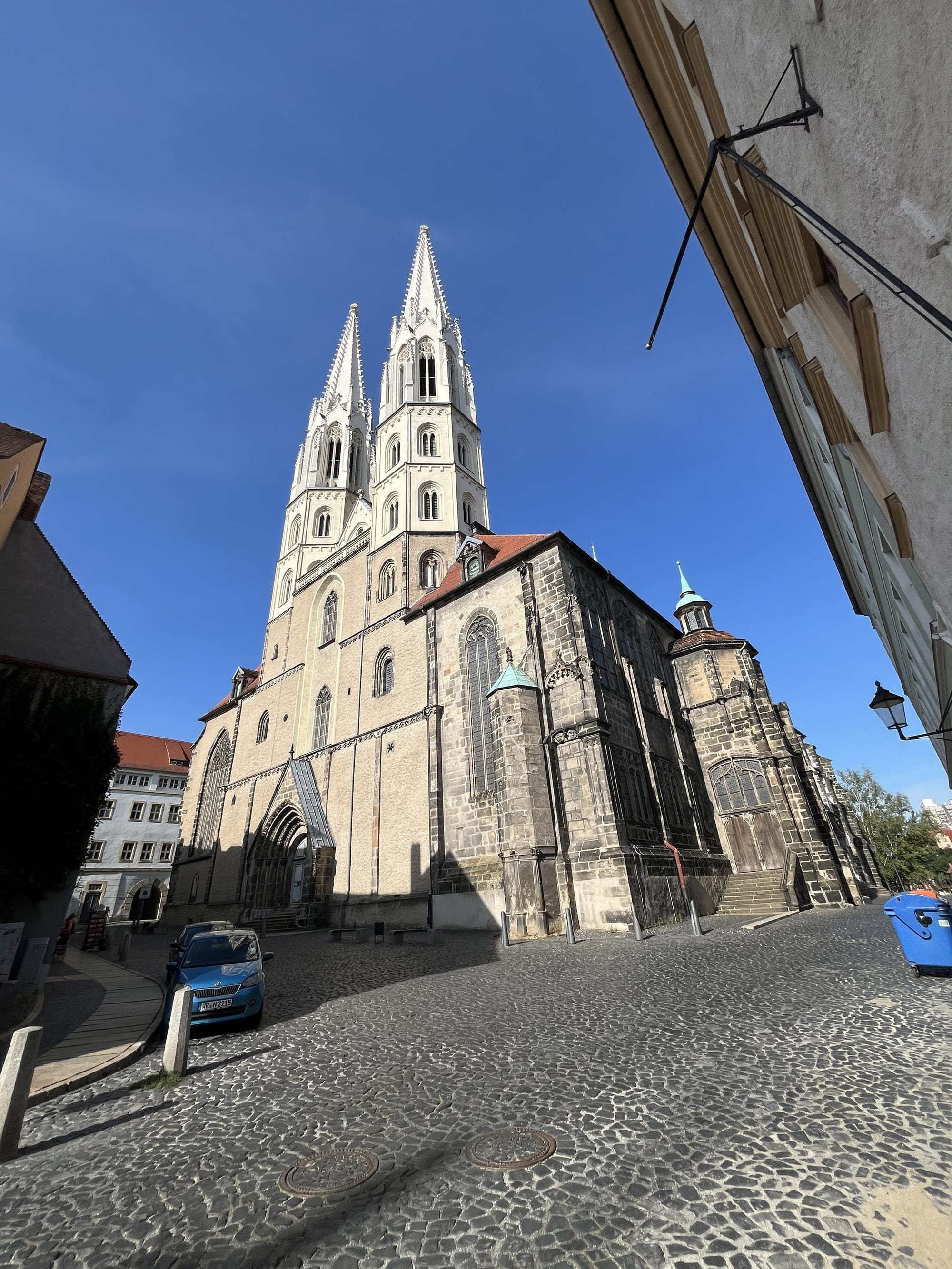 St Peter's Church in Görlitz, Germany, viewed from a cobbled street, with its twin Gothic spires rising above the historic city centre against a clear blue sky.