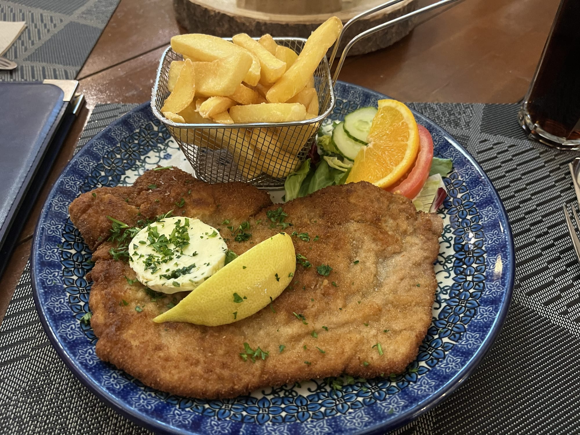 Large breaded schnitzel served on a patterned plate with chips, a lemon wedge, herb butter and a small side salad.