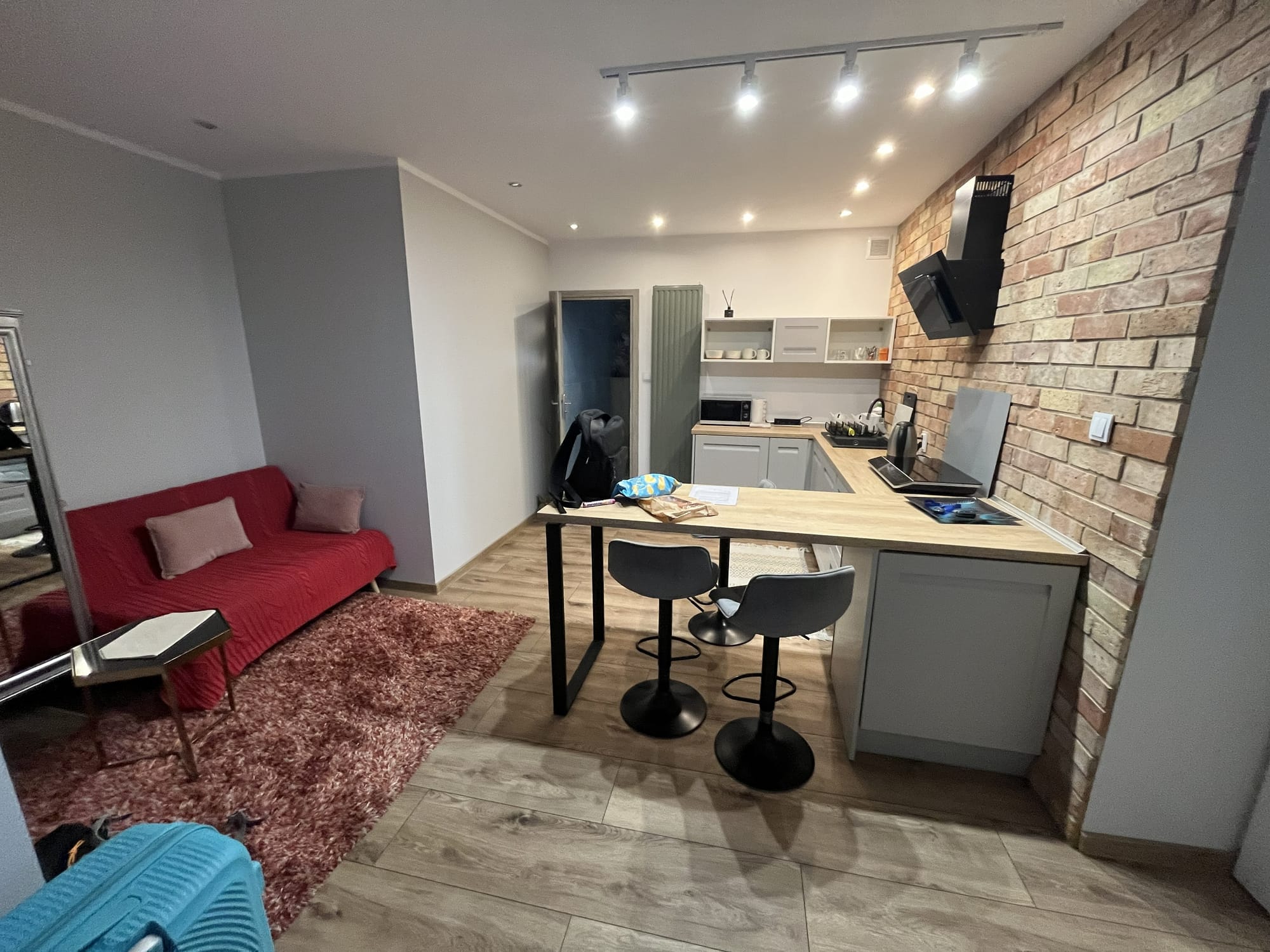 Modern apartment living area in Kraków near Borek Fałęcki station, featuring a compact kitchen with grey cabinetry, a breakfast bar with stools, a red sofa, exposed brick-effect wall and contemporary lighting