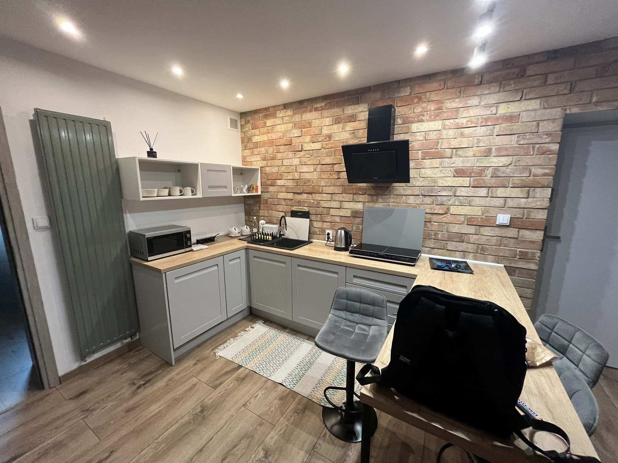 Modern apartment kitchen in Kraków near Borek Fałęcki station, featuring grey cabinets, wooden worktops, a dining table, microwave, kettle, induction hob and exposed brick-effect wall.