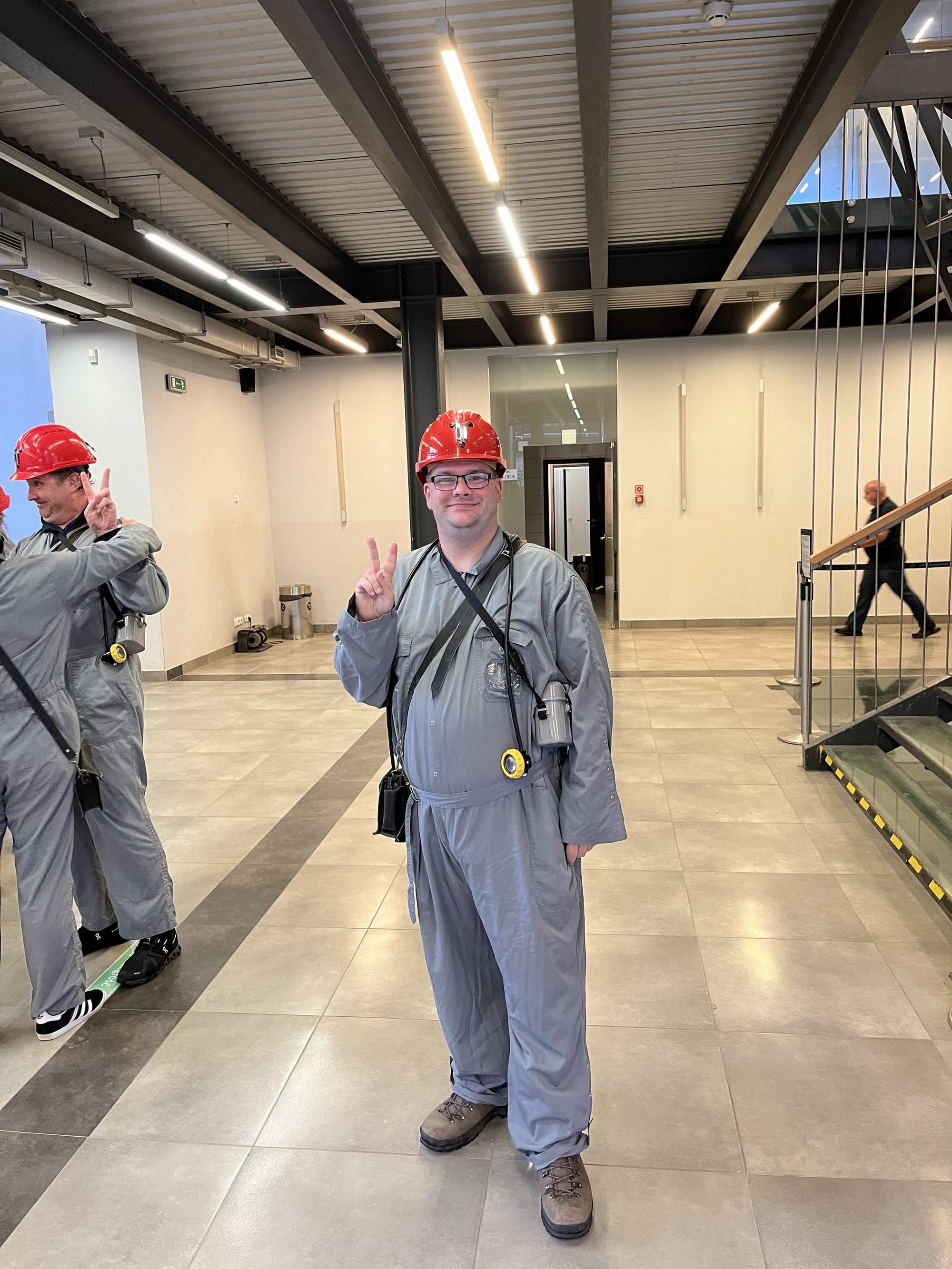 Visitor wearing a red safety helmet, grey protective overalls and a mining lamp stands inside the Wieliczka Salt Mine visitor centre, making a peace sign before an underground tour.