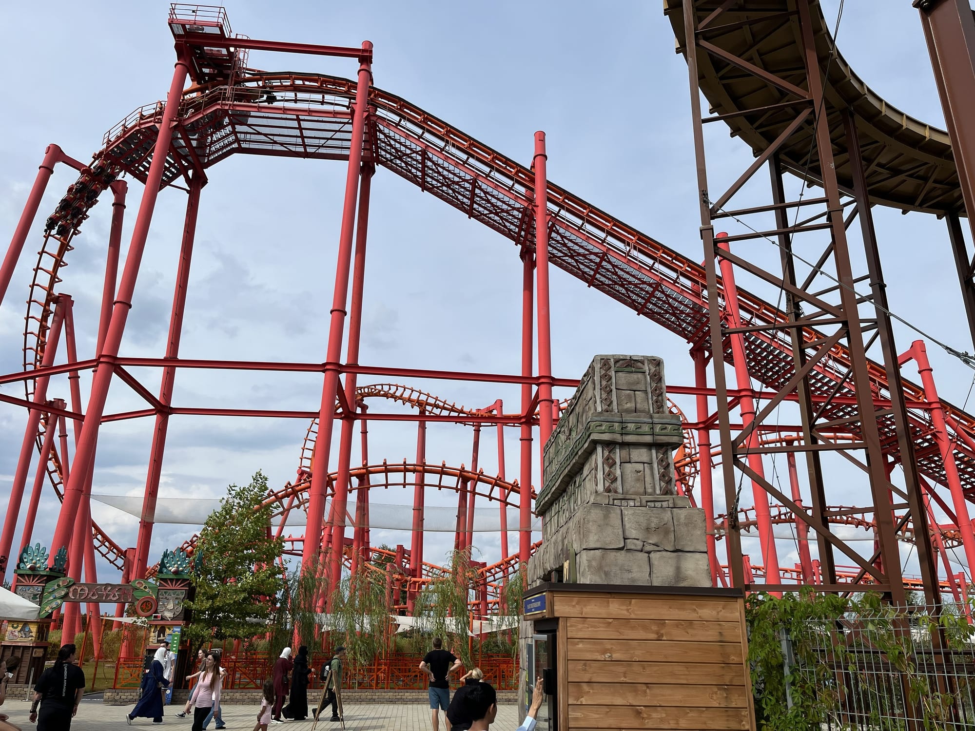 Mayan roller coaster at Energylandia, featuring bright red suspended track weaving through a dense network of twists, turns and support columns above the park's pathways.