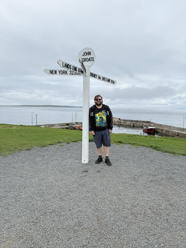 Person standing beside the John O' Groats signpost overlooking the harbour.