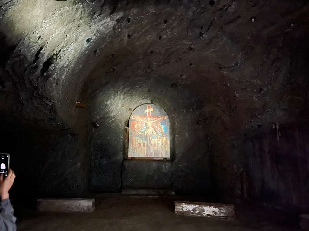 Religious artwork depicting the crucifixion displayed in a carved niche within an underground salt chamber at the Wieliczka Salt Mine, illuminated by a spotlight against dark rock walls.