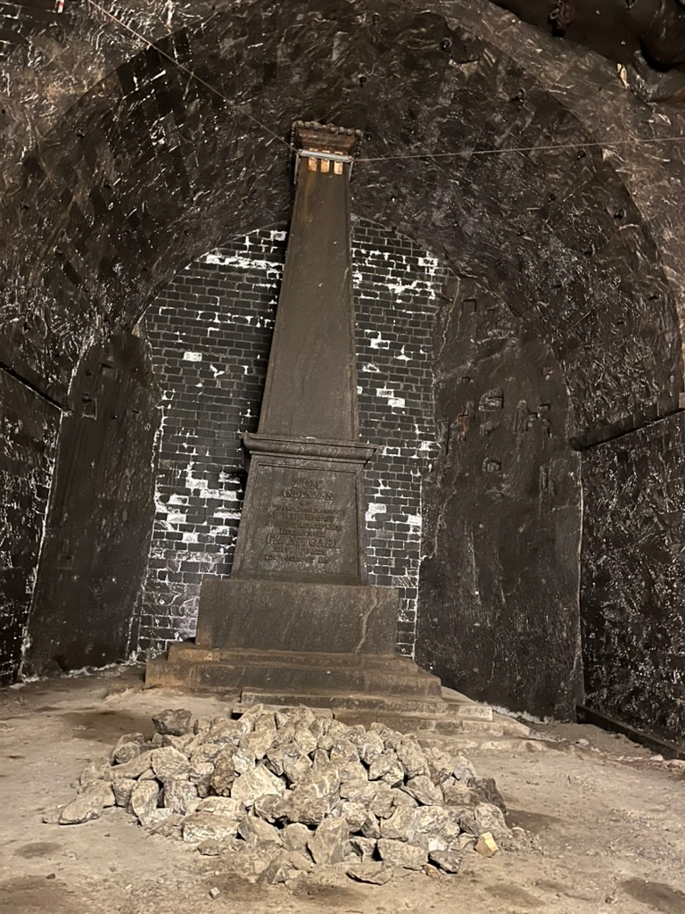 Stone obelisk monument standing in an underground chamber at the Wieliczka Salt Mine, surrounded by dark salt-rock walls and a pile of loose rocks in the foreground.