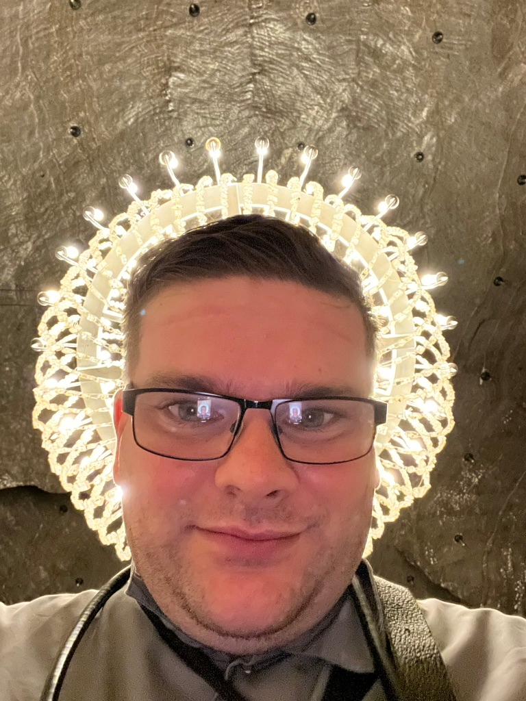Close-up selfie of a visitor wearing glasses inside the Wieliczka Salt Mine, standing beneath a large illuminated crystal chandelier that creates a halo-like effect against the salt-rock walls.