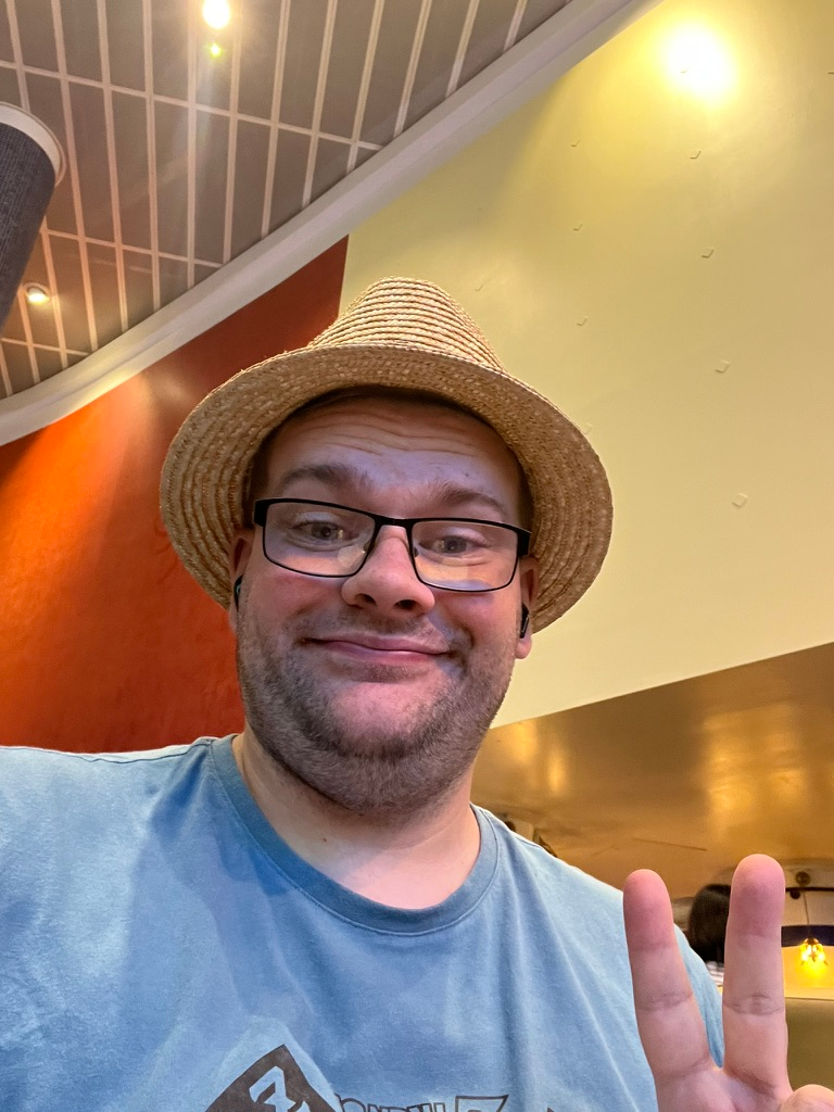 Close-up selfie of a person wearing glasses and a straw hat, making a peace sign while seated in a café area at Manchester Airport, with warm lighting and a curved ceiling overhead.