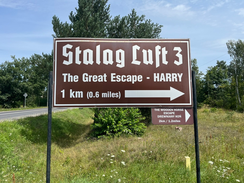 Brown roadside sign for Stalag Luft III pointing towards the Great Escape tunnel site known as “Harry”, located 1 kilometre away, with a second sign directing visitors to the Wooden Horse Escape site.