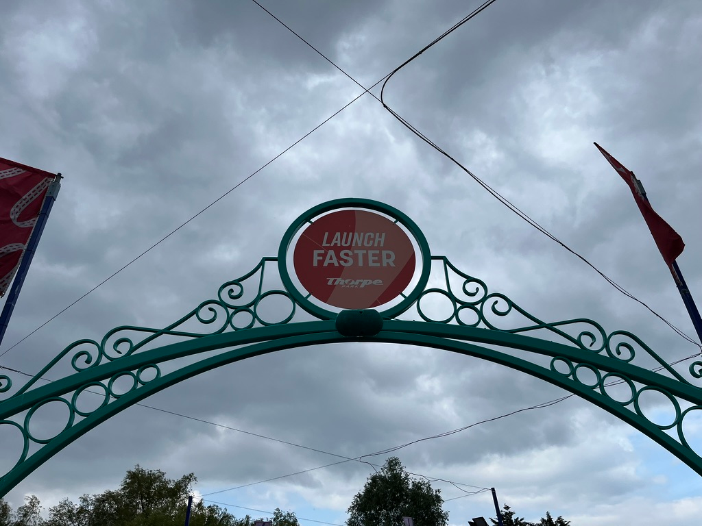 Decorative archway at Thorpe Park featuring a circular red sign reading “Launch Faster”, set against dramatic dark clouds with flags and overhead ride cables visible.