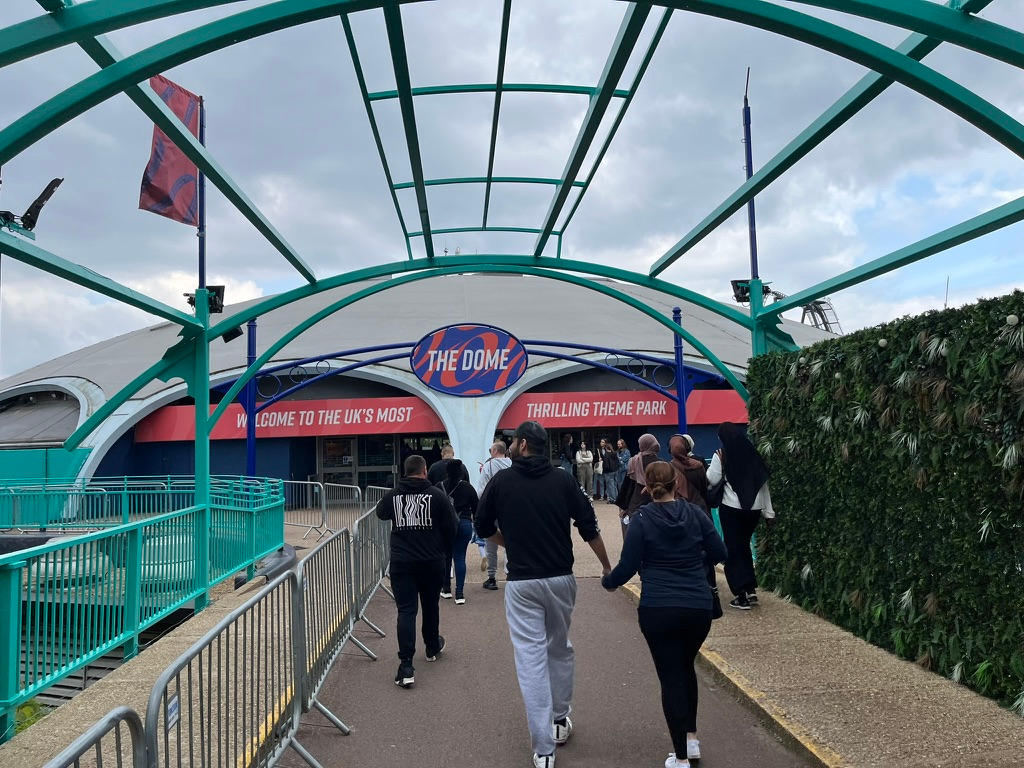 Visitors walk beneath a turquoise canopy towards The Dome entrance at Thorpe Park, with barriers lining the pathway and dramatic clouds overhead.