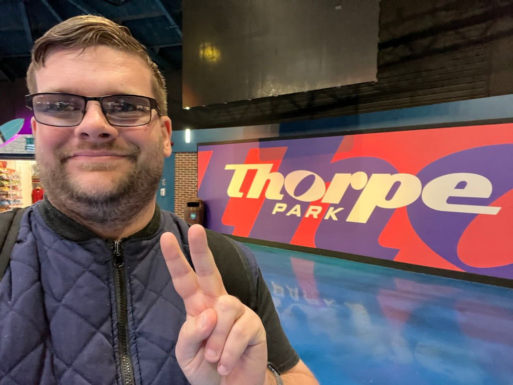 Visitor posing for a selfie inside Thorpe Park, making a peace sign in front of a large colourful Thorpe Park logo display within The Dome entrance complex.