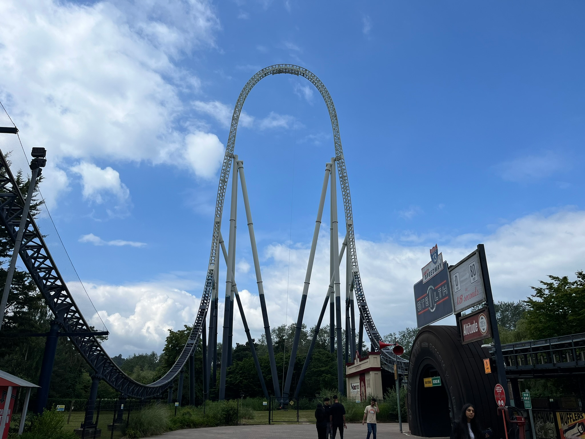Stealth roller coaster at Thorpe Park, featuring its towering top hat element rising 205 feet into the air, with the ride's entrance signage visible beneath a bright blue sky and scattered clouds.