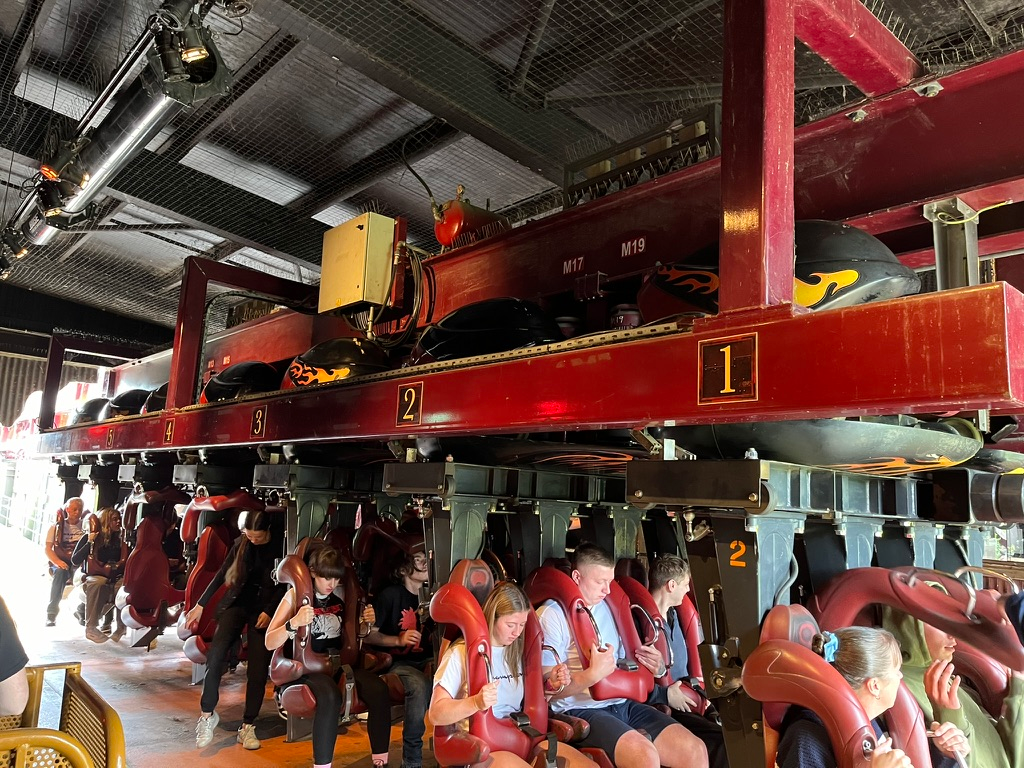 Riders seated on a Nemesis Inferno roller coaster train at Thorpe Park station, secured by over-the-shoulder restraints as the inverted coaster prepares to depart from the themed volcano-inspired loading area.