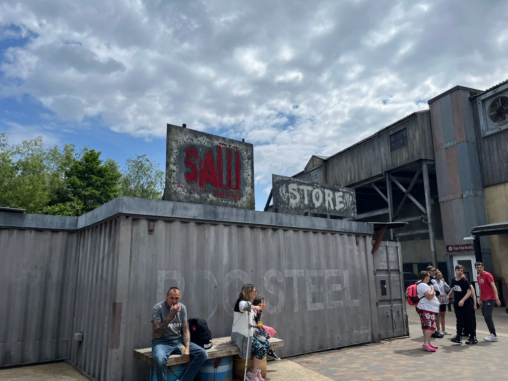 Entrance area for SAW – The Ride at Thorpe Park, featuring industrial-themed buildings, weathered signs reading “SAW” and “STORE”, shipping container-style structures and visitors gathered outside beneath a cloudy sky.