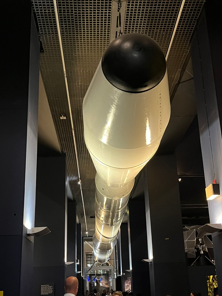 NASA Scout launch vehicle suspended from the ceiling of the Science Museum's Space gallery in London, viewed from below along its full length, with exhibition displays and visitors beneath the illuminated rocket.