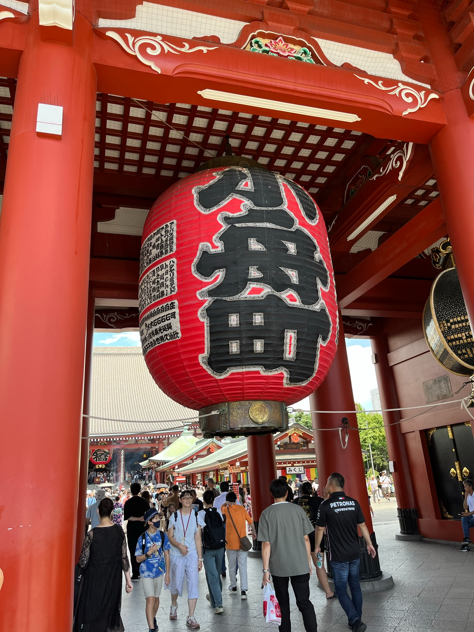 The giant red Kaminarimon lantern hangs beneath vermilion temple gates at Sensō-ji in Asakusa, with visitors passing beneath and temple buildings visible beyond.
