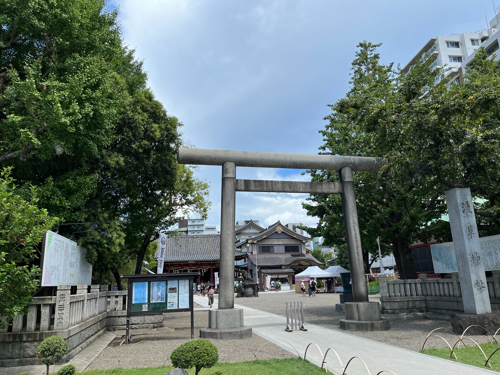 Stone torii gate at Asakusa Shrine in Tokyo, with shrine buildings, trees and visitors visible beyond the entrance under a partly cloudy summer sky.