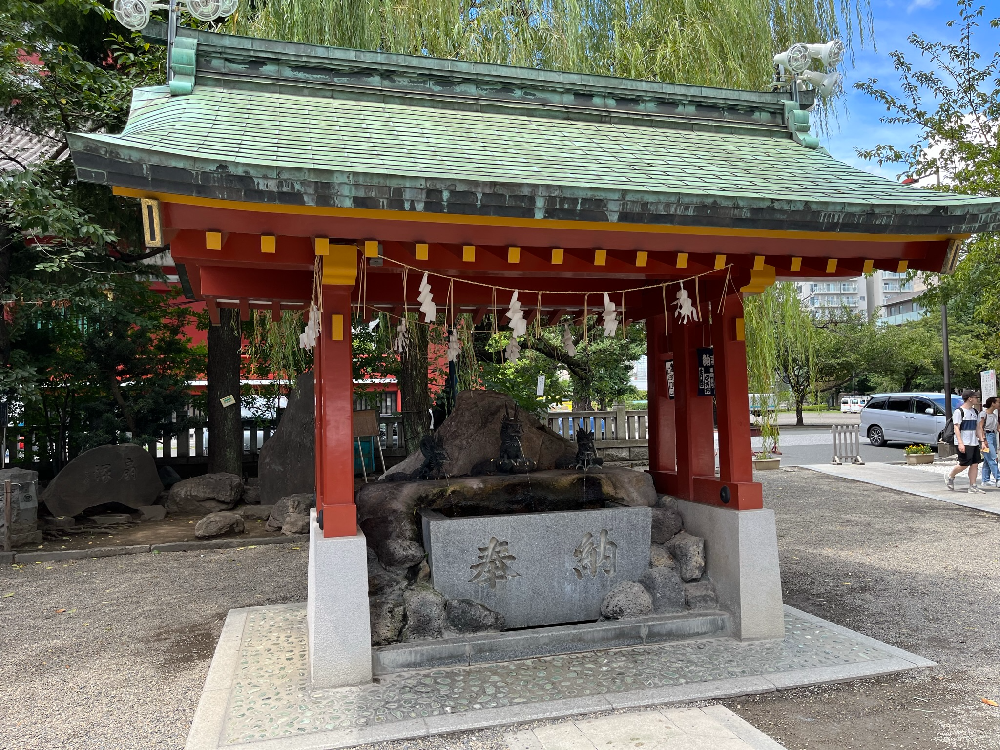 Temizuya water purification pavilion at Asakusa Shrine, featuring a red wooden structure, stone water basin and decorative dragon fountains beneath leafy trees.