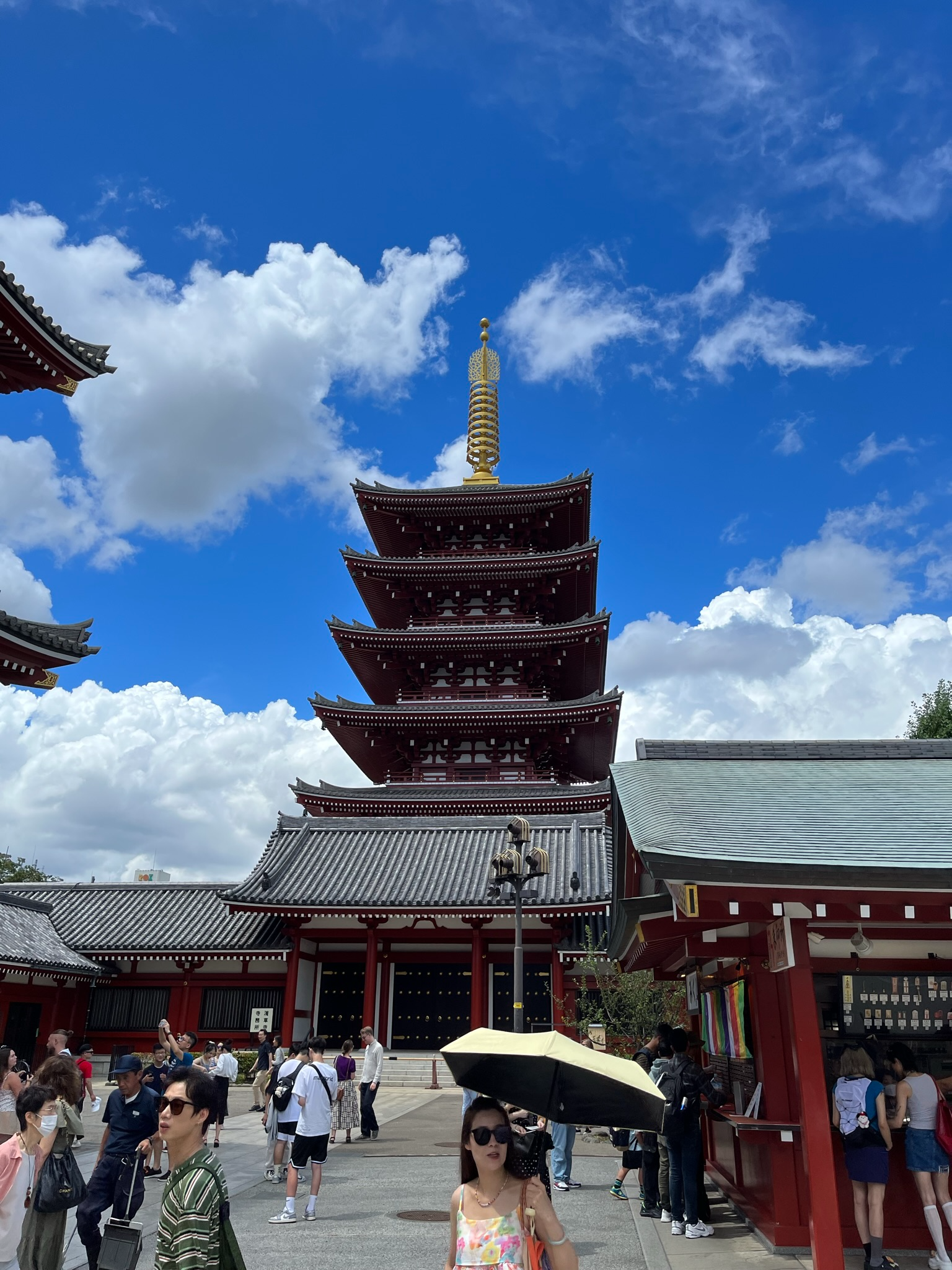 Five-storey pagoda at Sensō-ji Temple in Asakusa, framed by temple buildings and visitors beneath bright blue skies and towering white summer clouds.