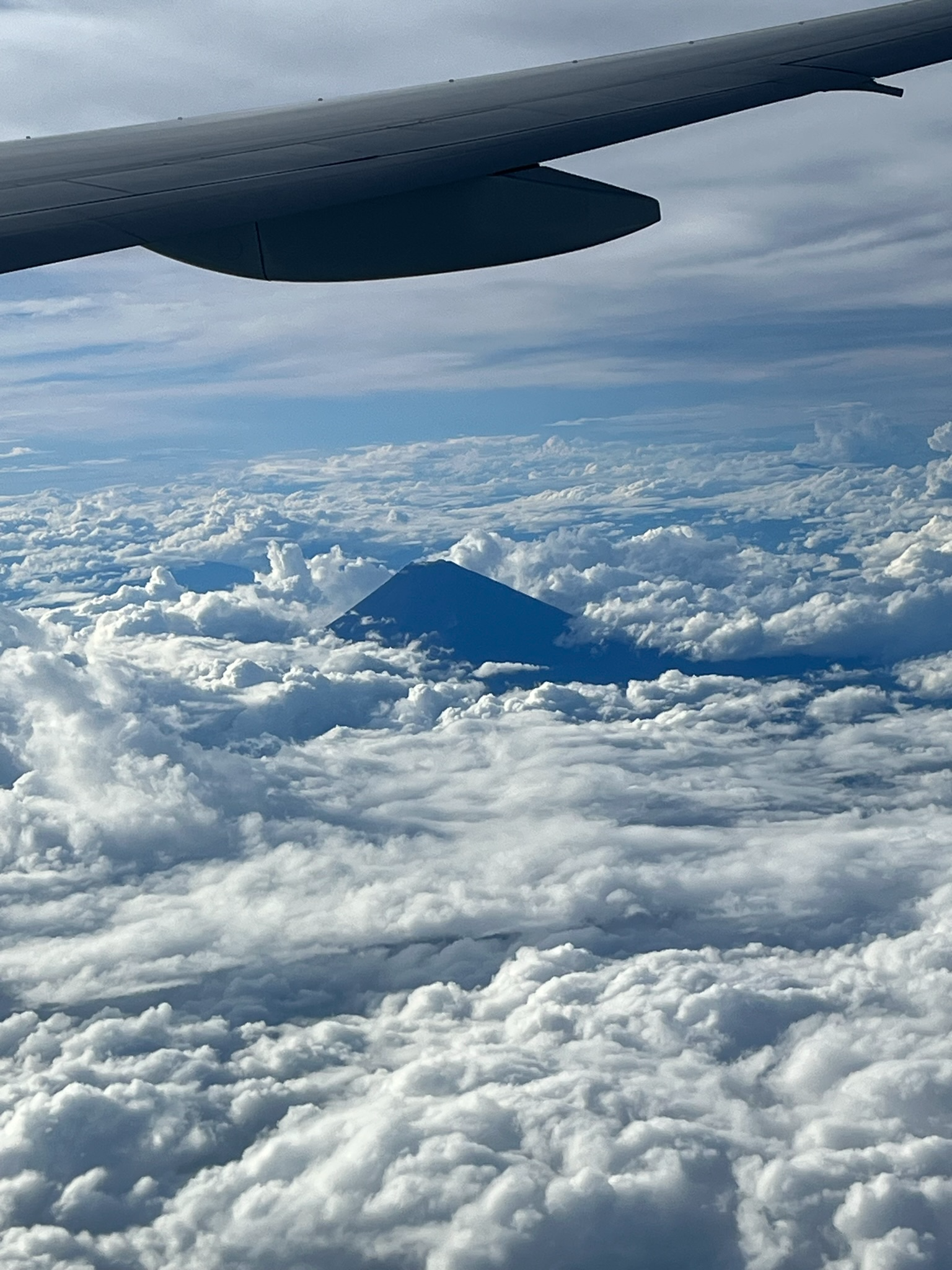 View of Mount Fuji from an airplane above the clouds, with the mountain's distinctive cone-shaped silhouette visible beneath an aircraft wing high over Japan.
