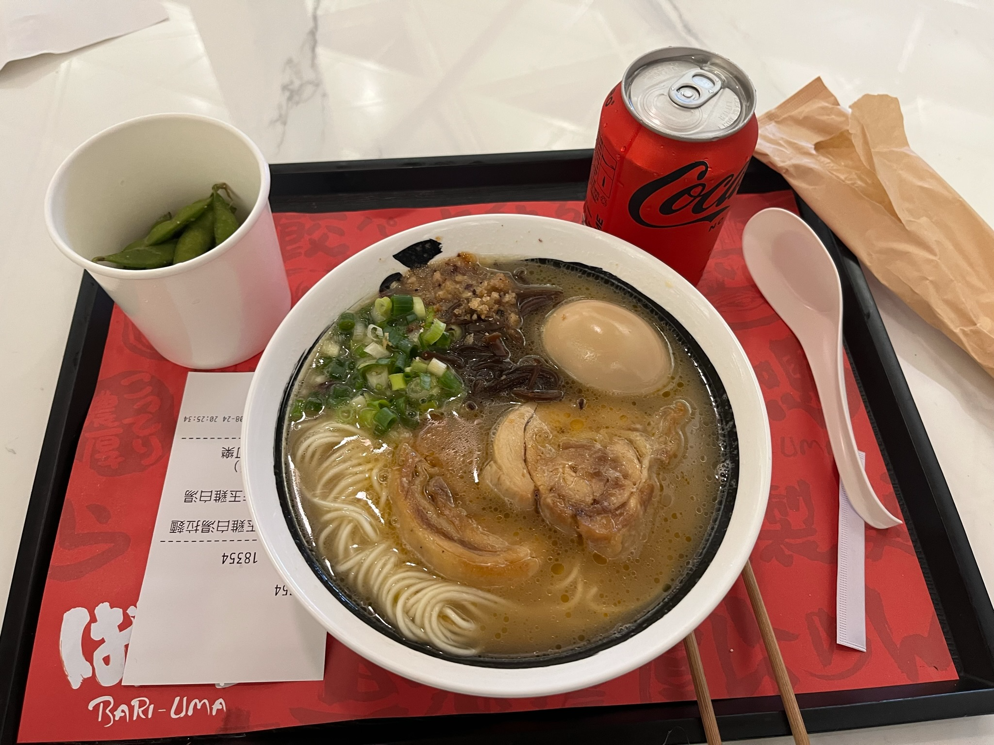 Bowl of tonkotsu ramen with chashu pork, seasoned egg and spring onions at Bari-Uma in Hong Kong International Airport, served with edamame and a can of Coca-Cola.