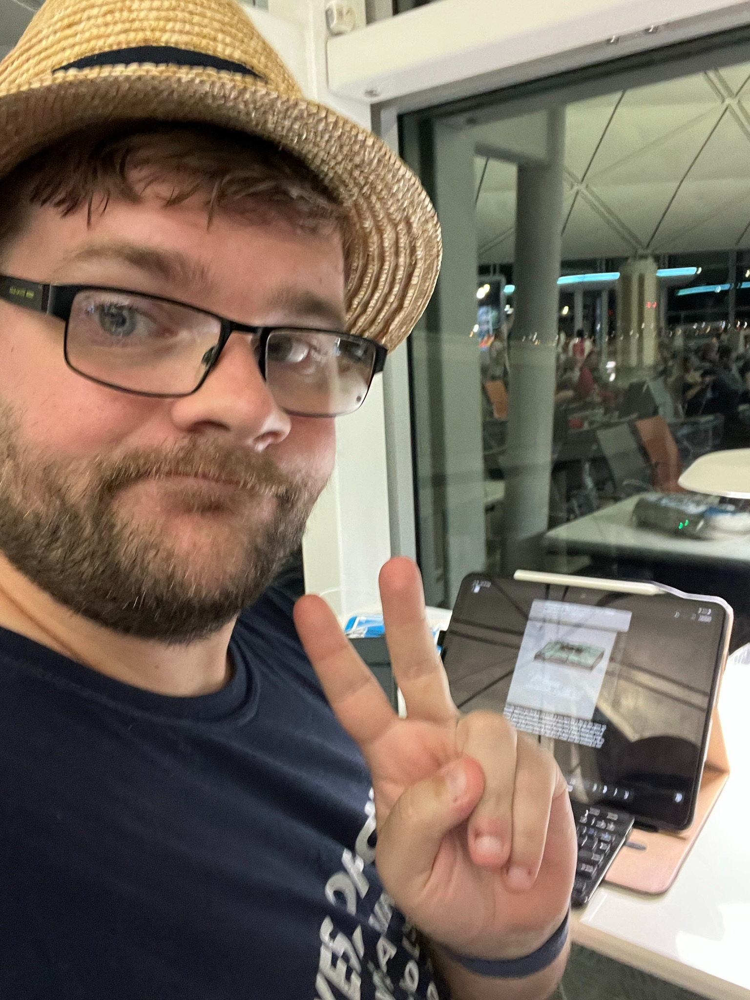 Traveller wearing a straw hat and glasses works on a travel blog using a tablet and keyboard at Hong Kong International Airport, while waiting near the departure gates.