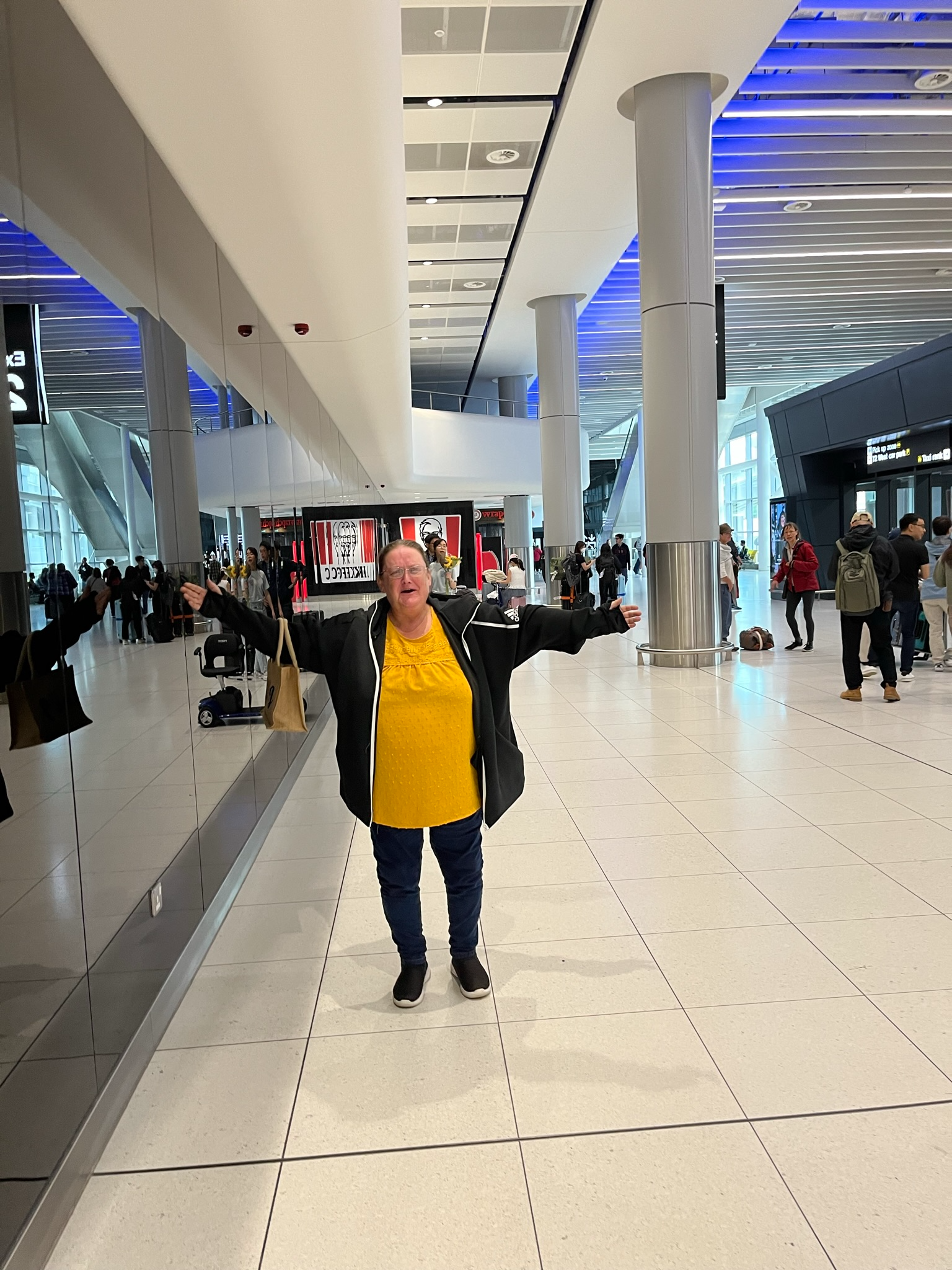 Person standing in an airport arrivals hall with arms outstretched, waiting to welcome a returning traveller home amid the bright, modern terminal surroundings.