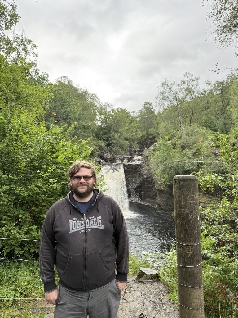 Person standing in front of the Falls of Falloch waterfall in woodland.