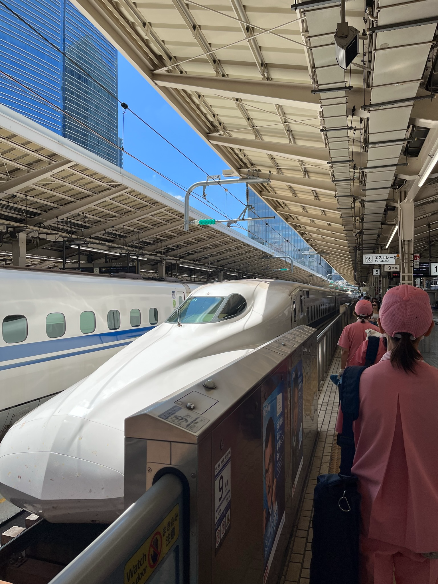 N700 Series Shinkansen bullet train at a station platform, with its streamlined nose beside waiting passengers under a large covered concourse and clear blue skies.