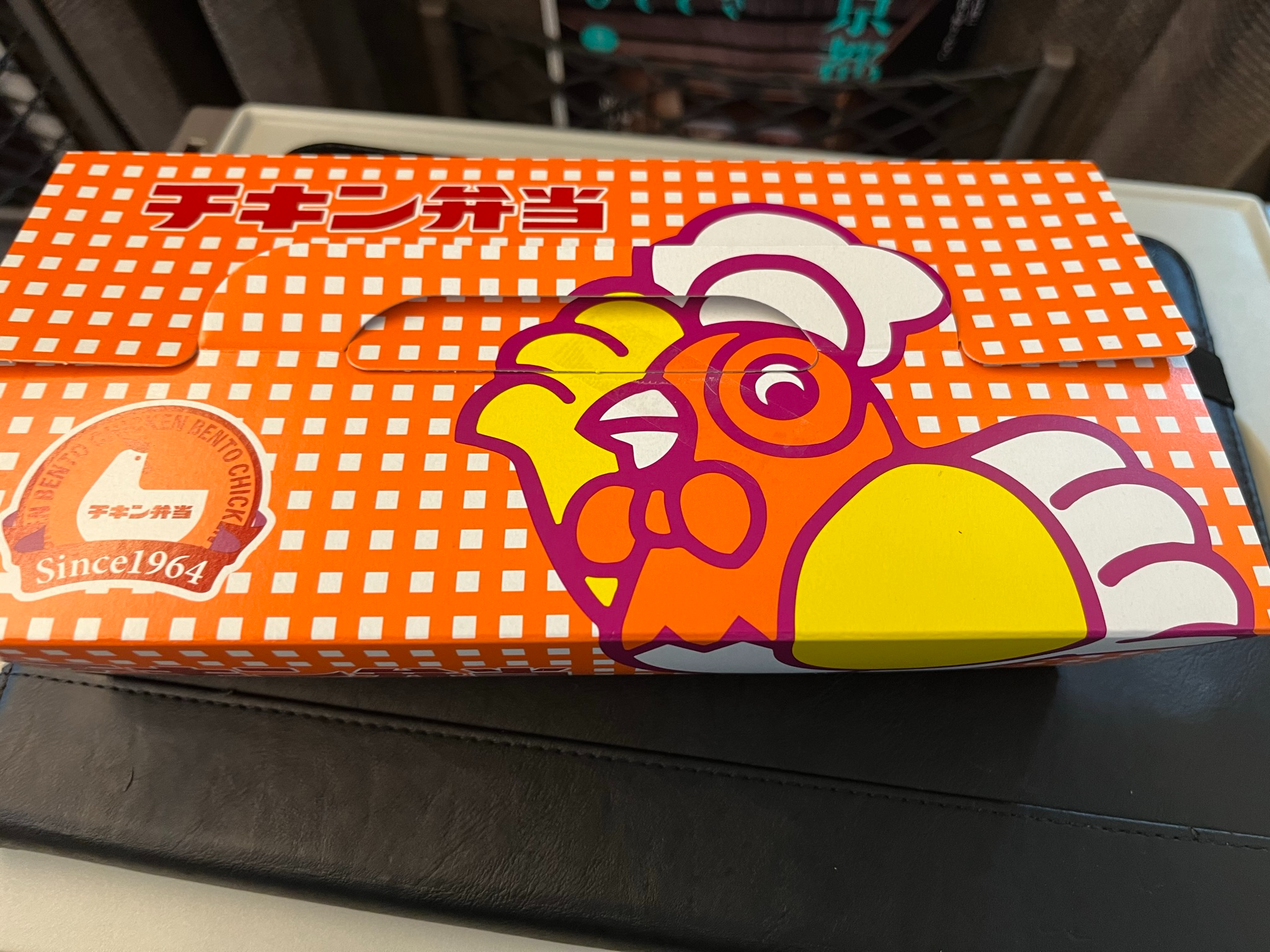 Boxed chicken bento ekiben on a Shinkansen tray table, featuring a bright orange and white package with a cartoon chicken and Japanese text dating the product to 1964.