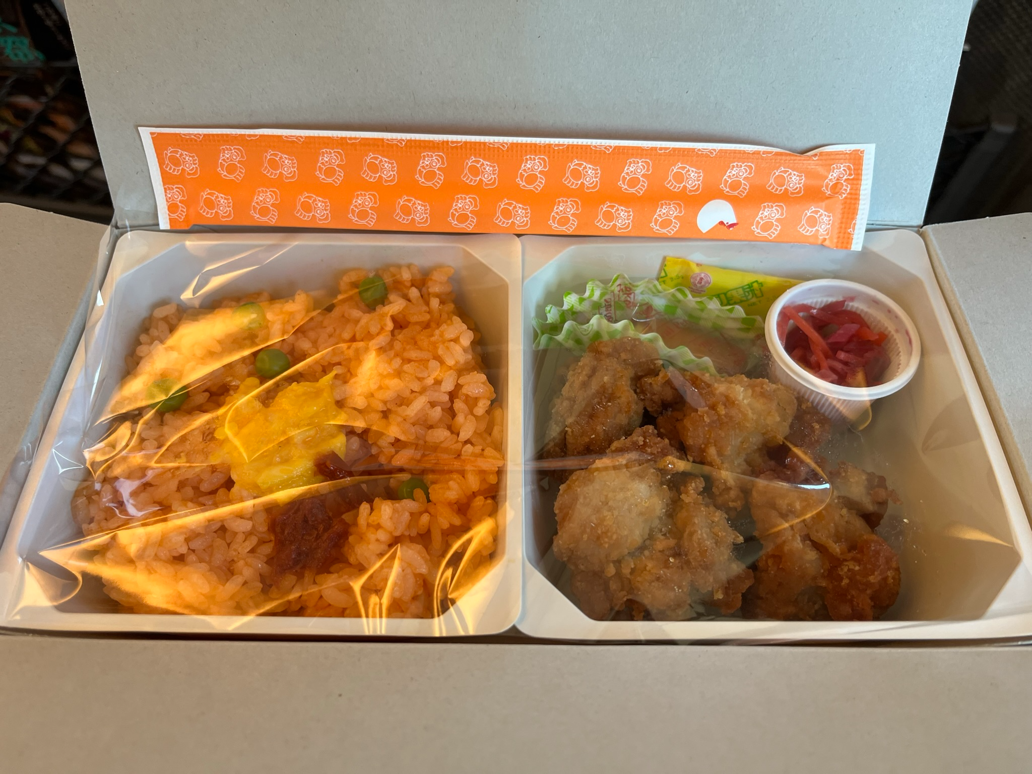 Opened chicken ekiben on a Shinkansen tray table, featuring seasoned rice with peas and egg alongside pieces of fried chicken, pickles and condiments in separate compartments.