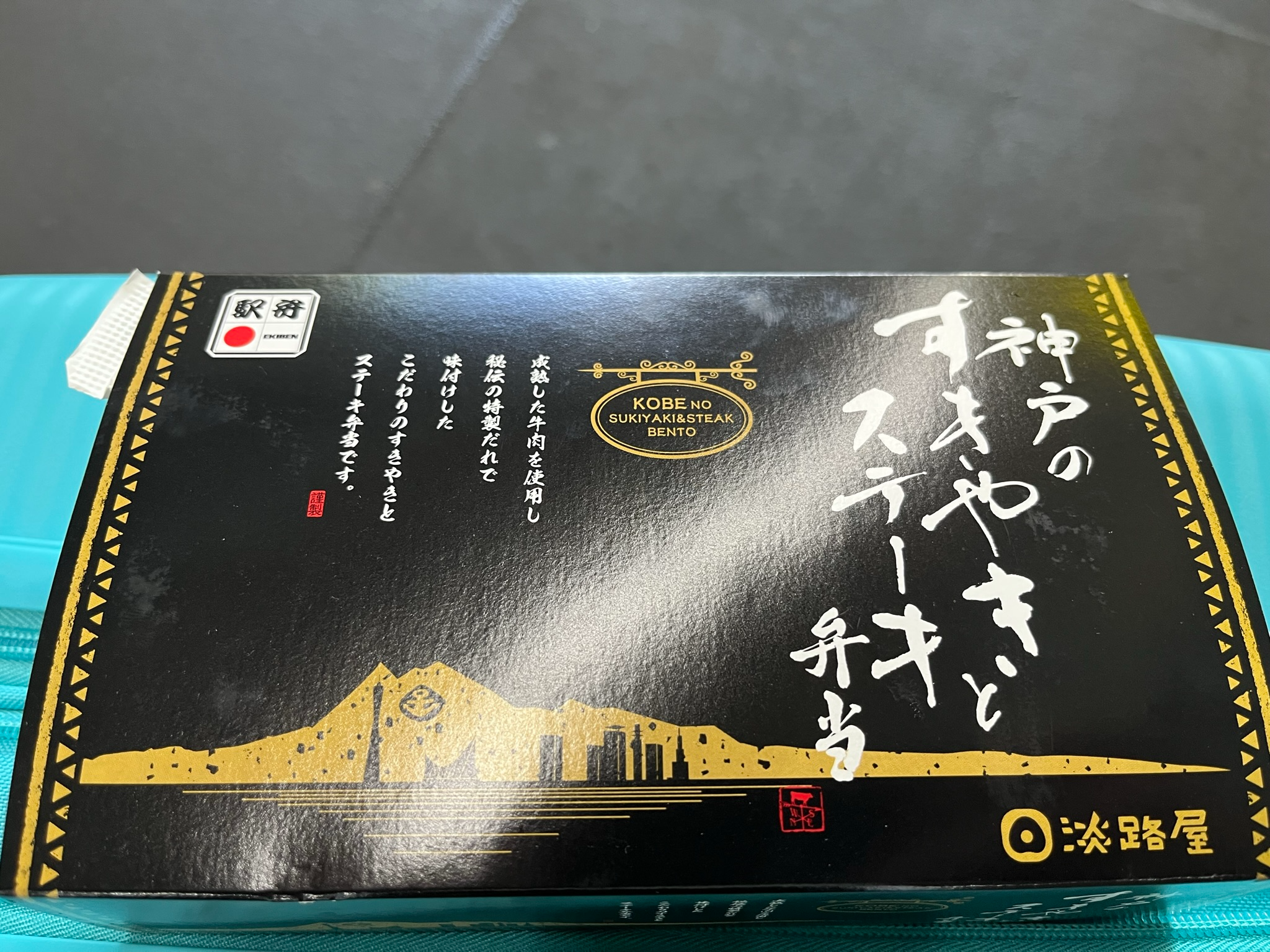 Box of Kobe Sukiyaki & Steak Ekiben on a train tray table, featuring black and gold packaging with Japanese calligraphy, a city skyline illustration and ekiben branding.