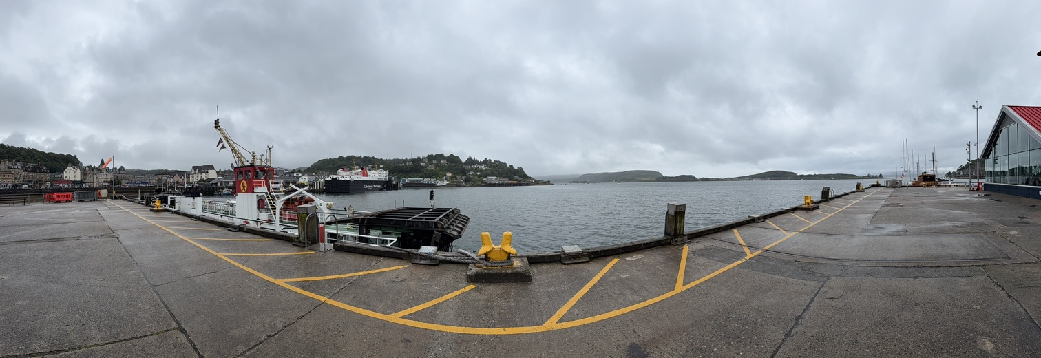 Panoramic view of Oban harbour with ferries and waterfront buildings under grey skies.