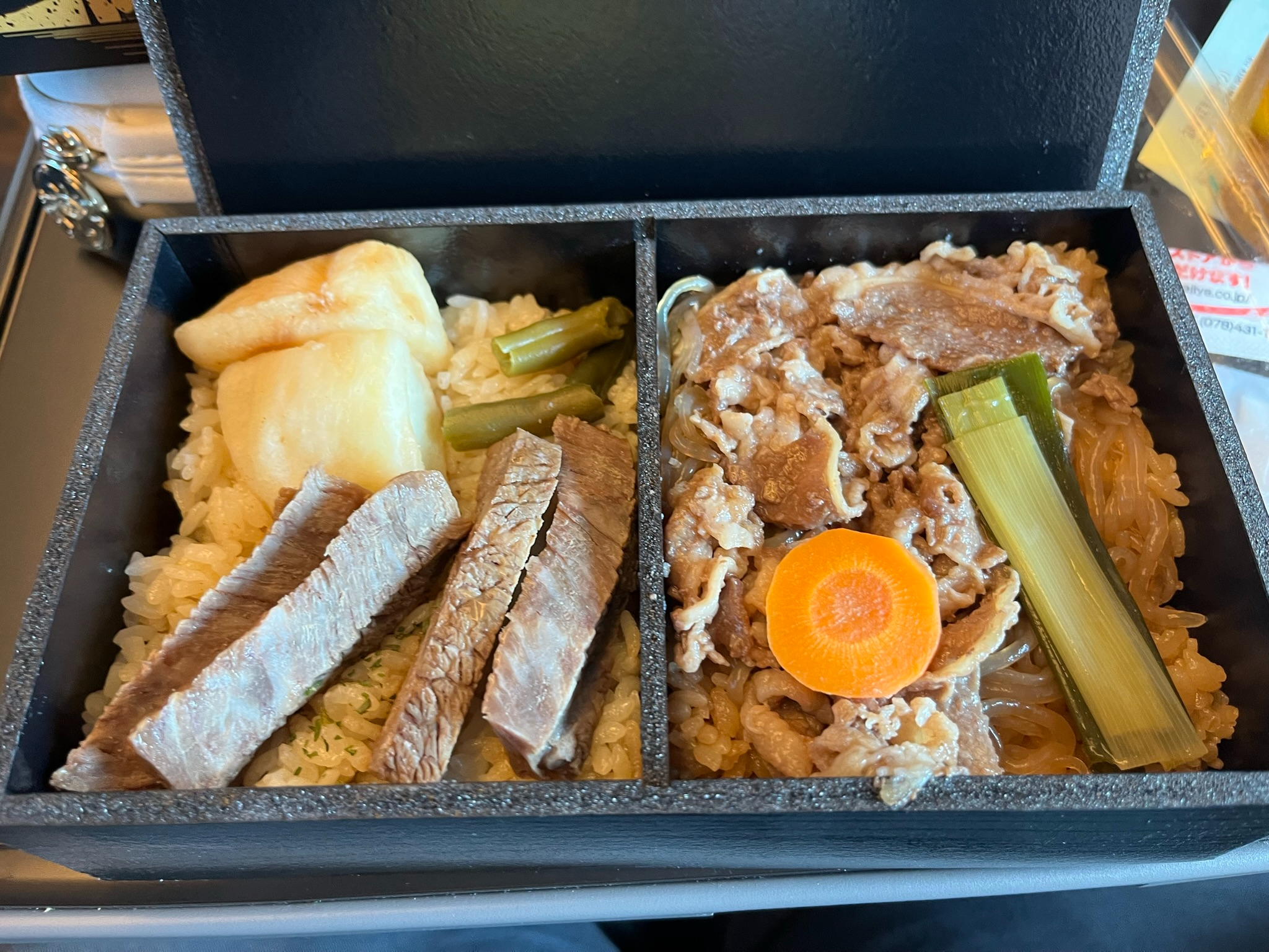 Opened Kobe Sukiyaki & Steak Ekiben featuring sliced steak, sukiyaki beef, rice, vegetables and pickles arranged in a black compartmentalised bento box on a train tray table.