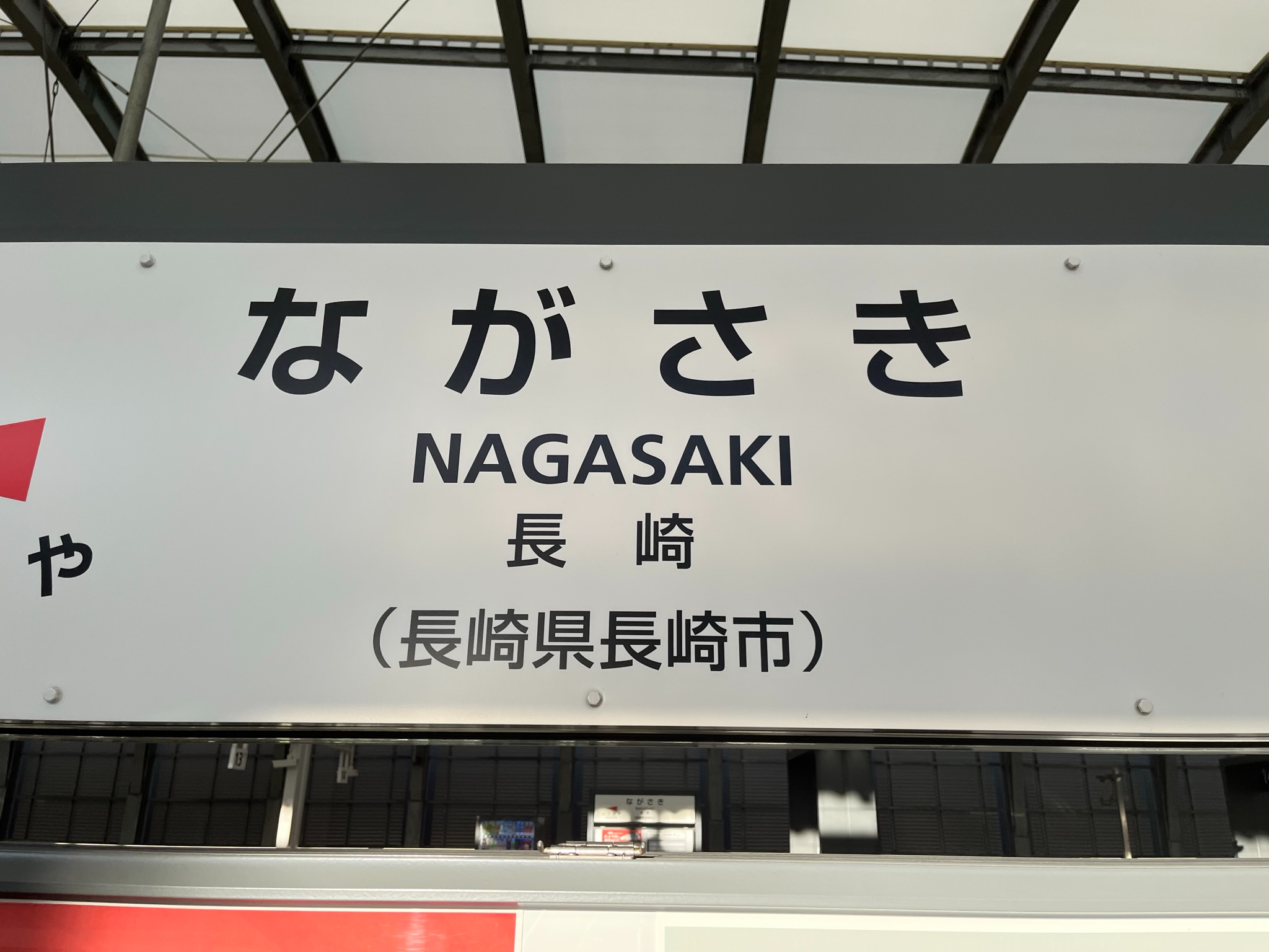 Nagasaki Station sign displaying the city name in Japanese and English, mounted above a railway platform inside the station with structural beams visible overhead.