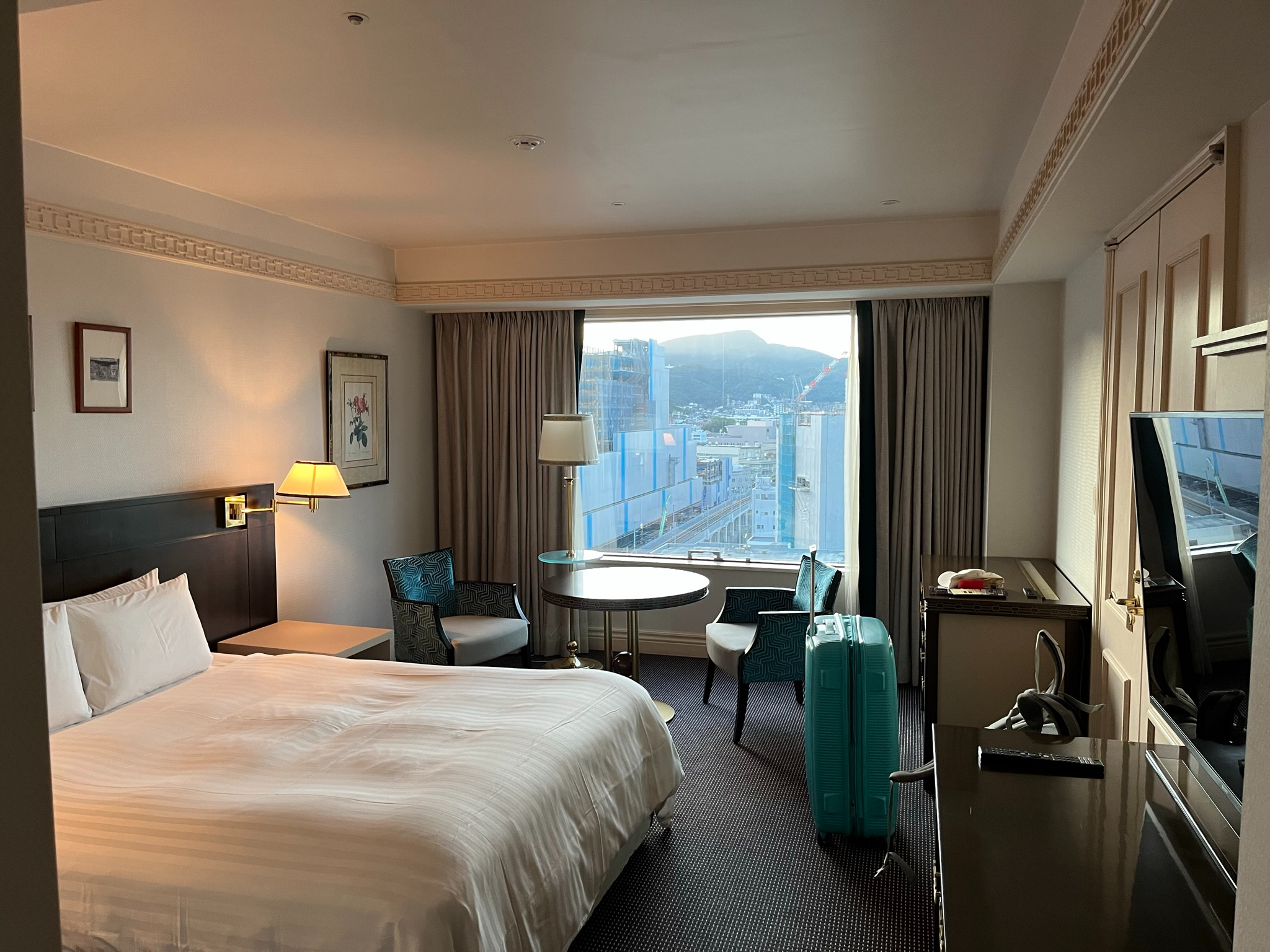Guest room at The Global View Nagasaki featuring a large bed, seating area, desk, television and a wide window overlooking the city and surrounding hills.