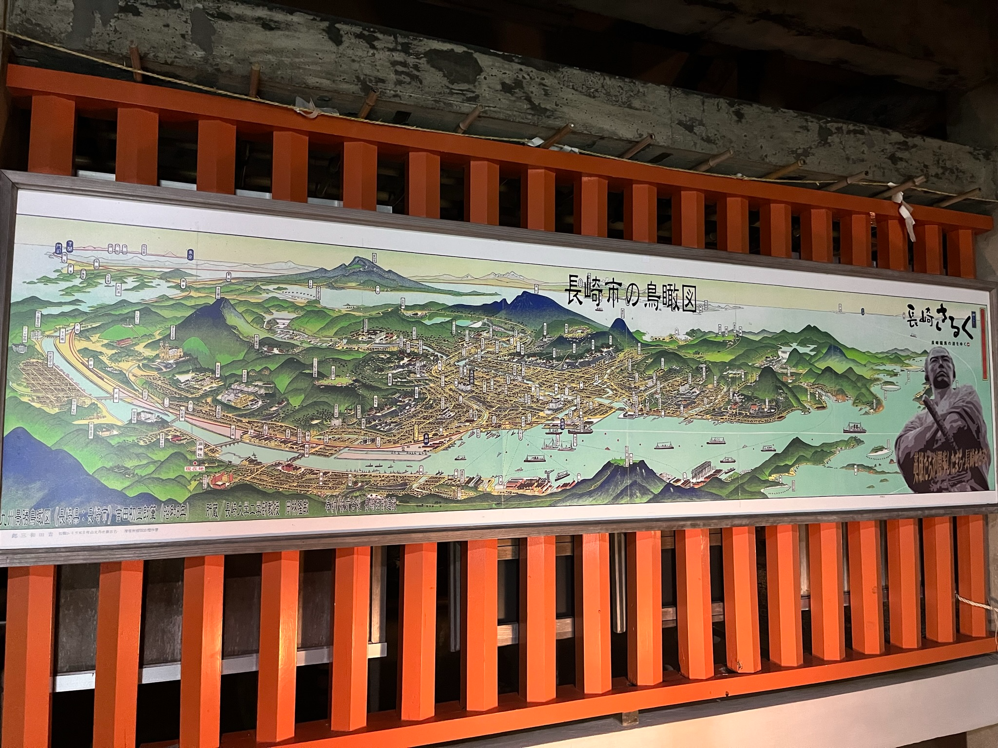 Illustrated panoramic map of Nagasaki displayed at Mount Inasa, showing the city's harbour, districts, mountains, bridges and notable landmarks across the surrounding landscape.