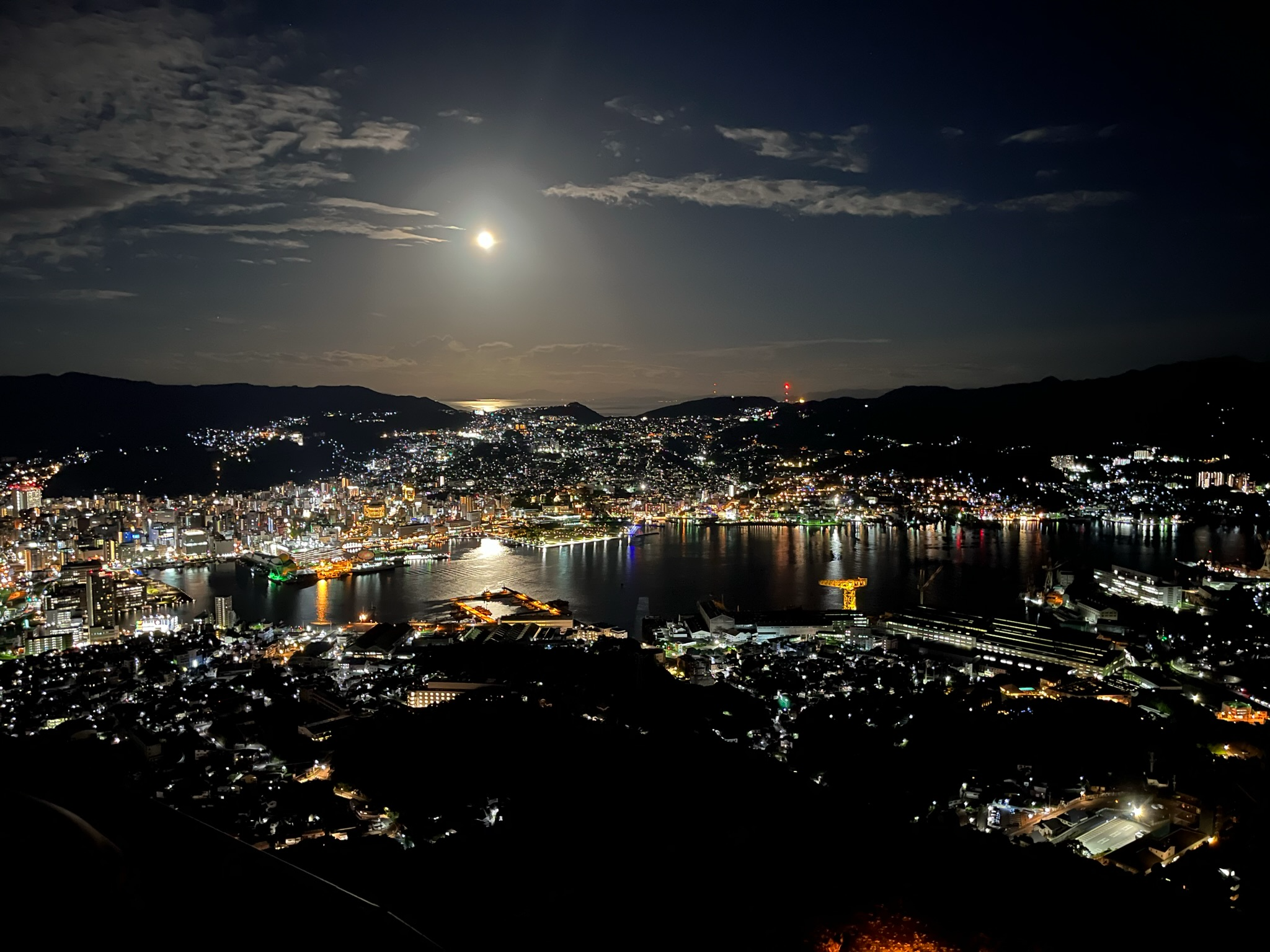 Night view from Mount Inasa overlooking Nagasaki Harbour, with city lights reflecting on the water, surrounding hills illuminated and the moon shining above the bay.