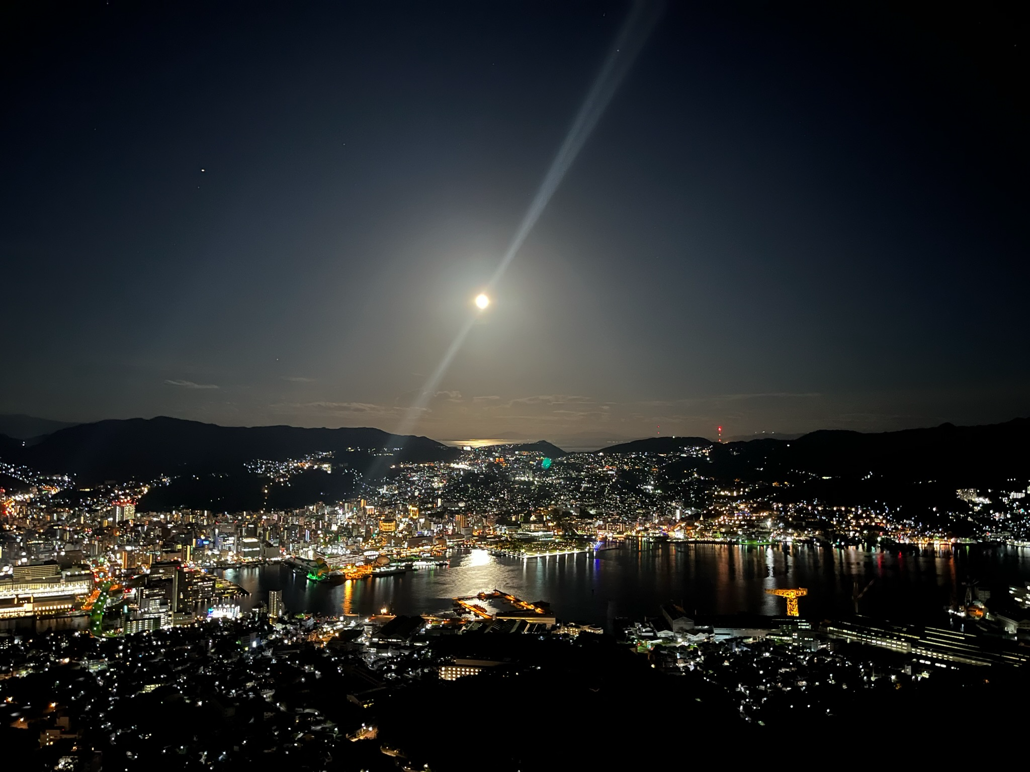 Night view from Mount Inasa across Nagasaki Harbour, with city lights stretching over hills and waterfront districts beneath a bright moon reflected on the distant sea.