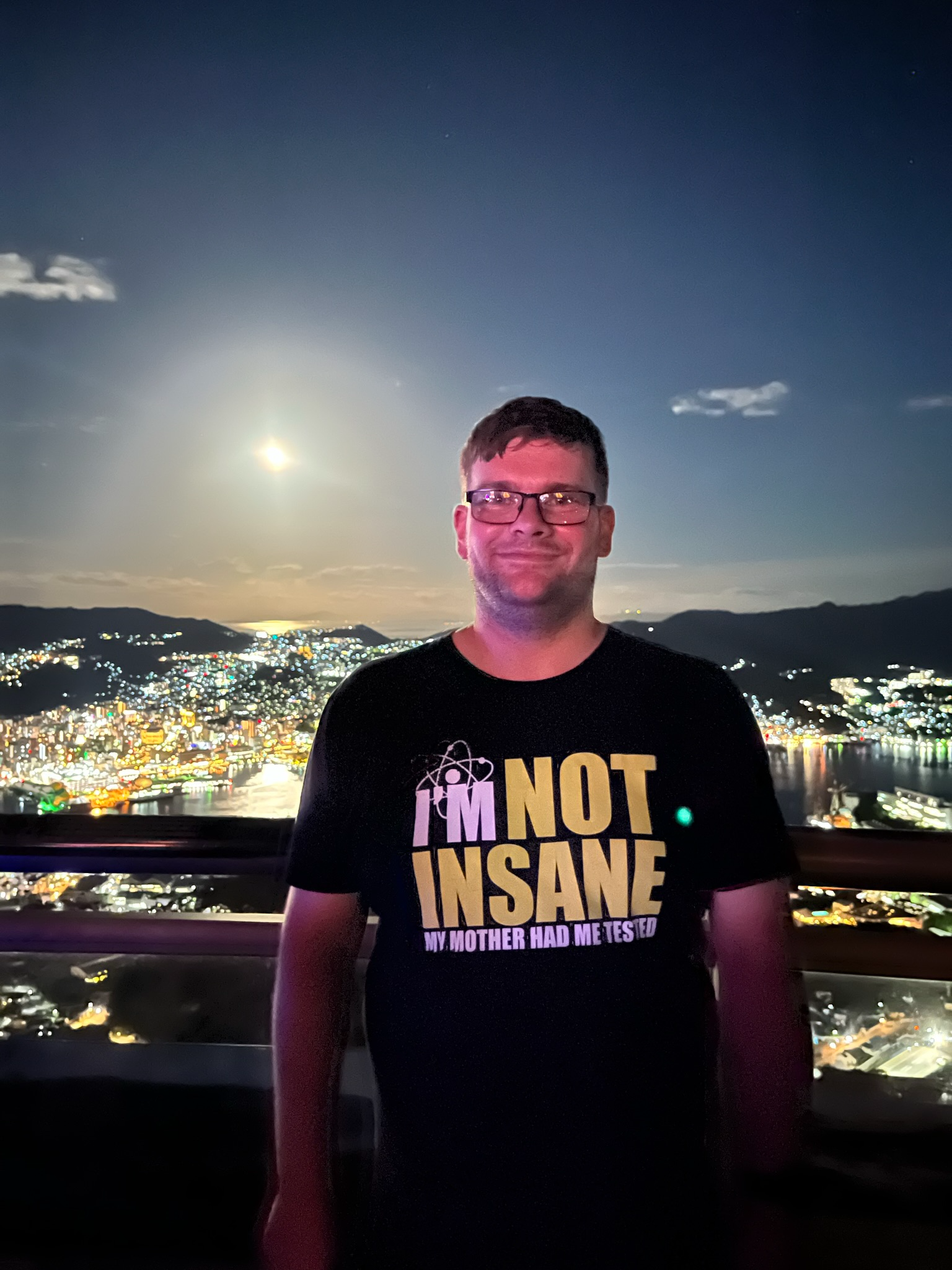 Visitor standing at the Mount Inasa observation deck overlooking Nagasaki at night, with city lights, the harbour and a bright moon illuminating the skyline beyond.