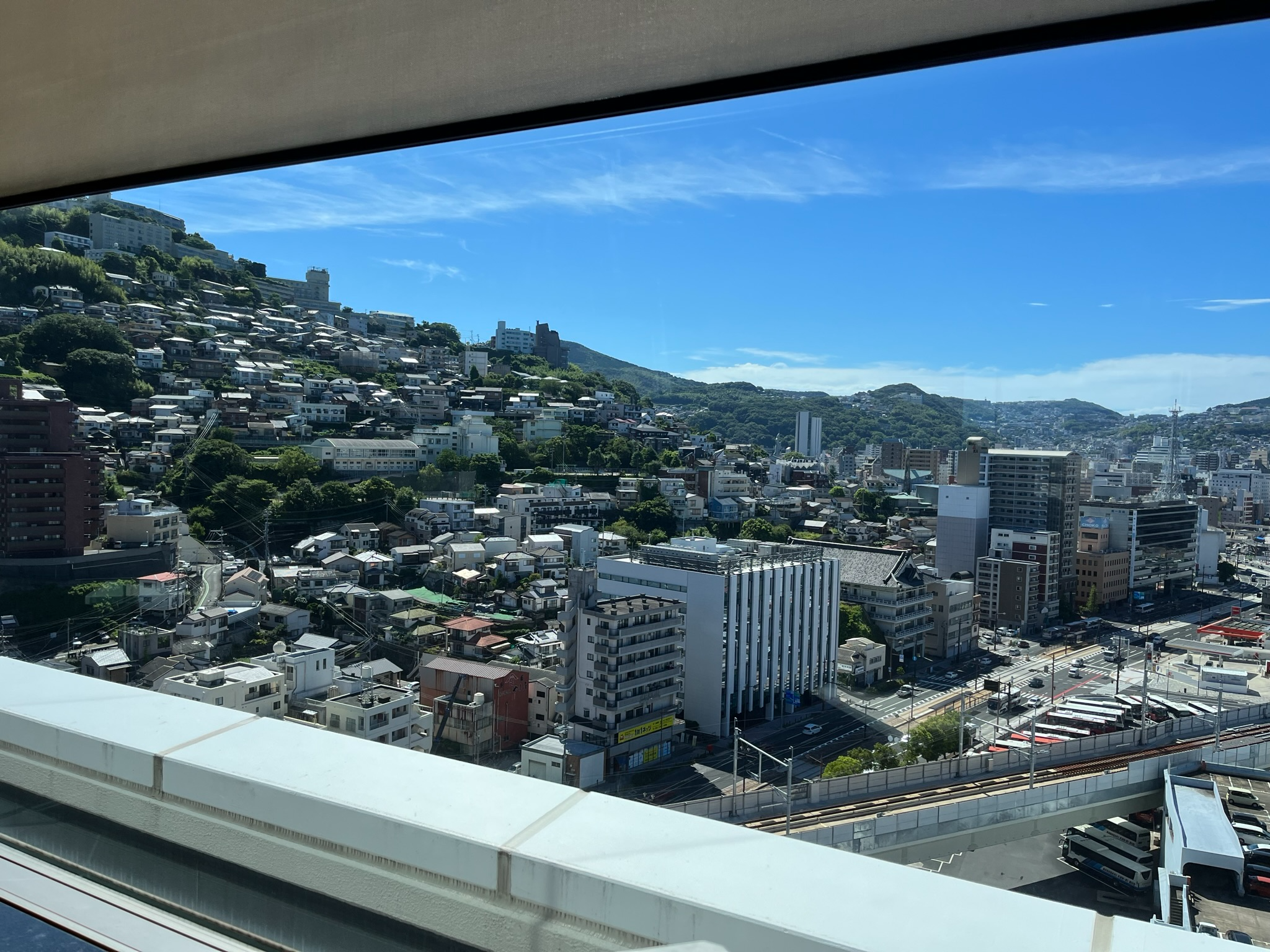 View across Nagasaki from a high vantage point, showing hillside homes, city buildings, roads and green mountains beneath clear blue skies on a sunny day.