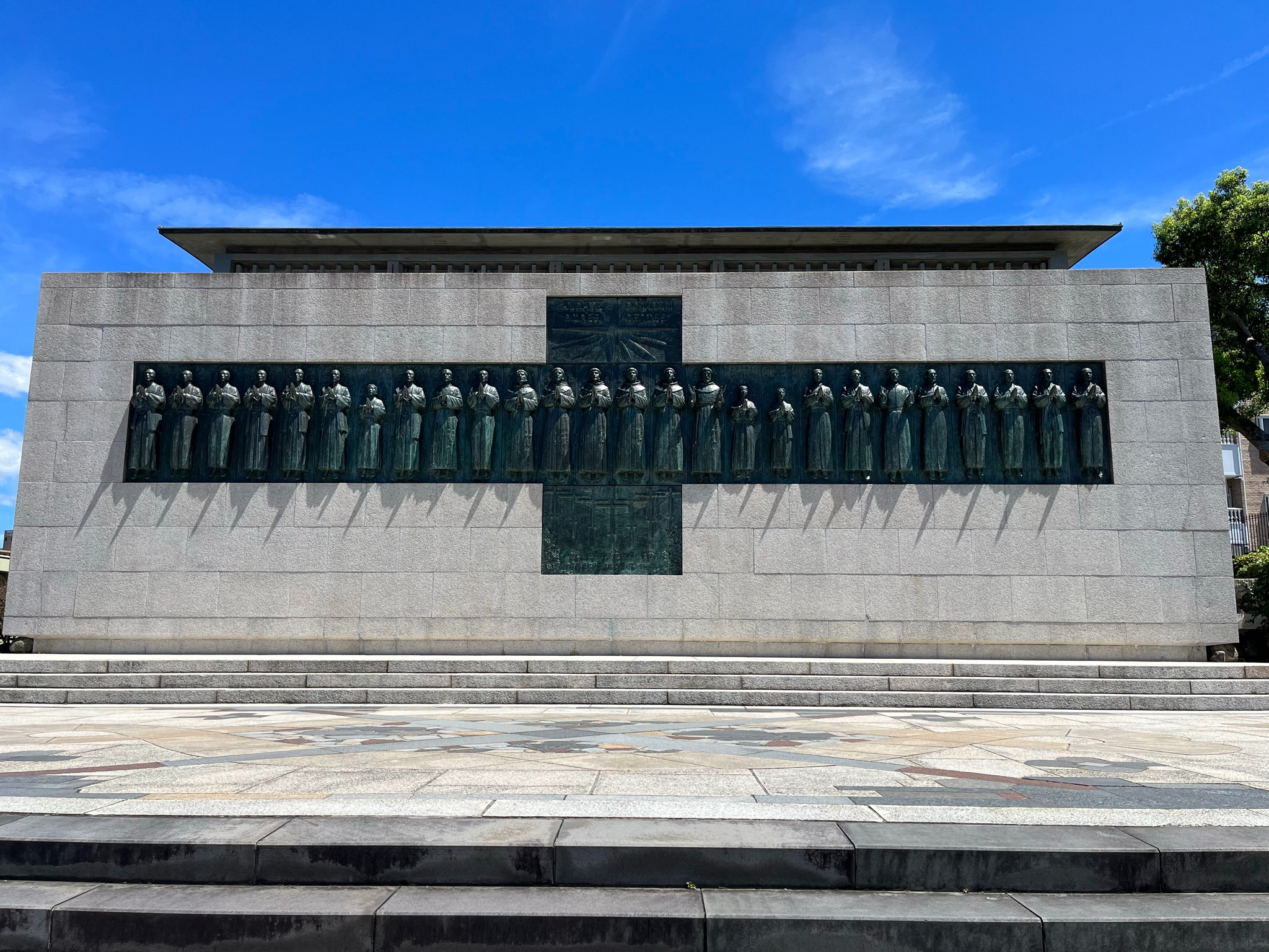 The Twenty-Six Martyrs Monument in Nagasaki, featuring bronze statues of the martyrs mounted on a large stone wall beneath a clear blue sky and bright sunshine.