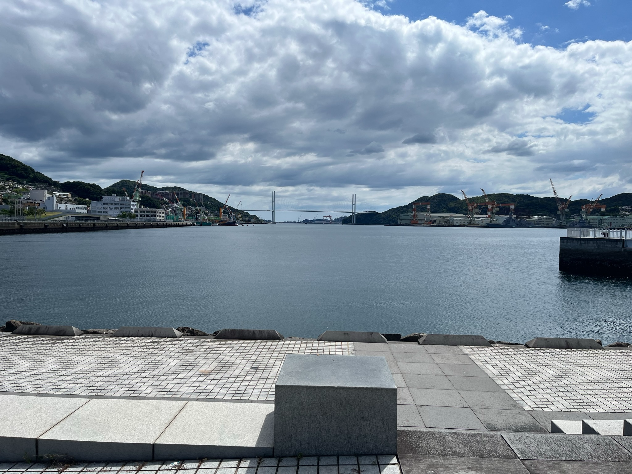 View across Nagasaki Harbour with calm water, shipyard cranes, waterfront facilities and distant hills beneath dramatic clouds and patches of blue sky.