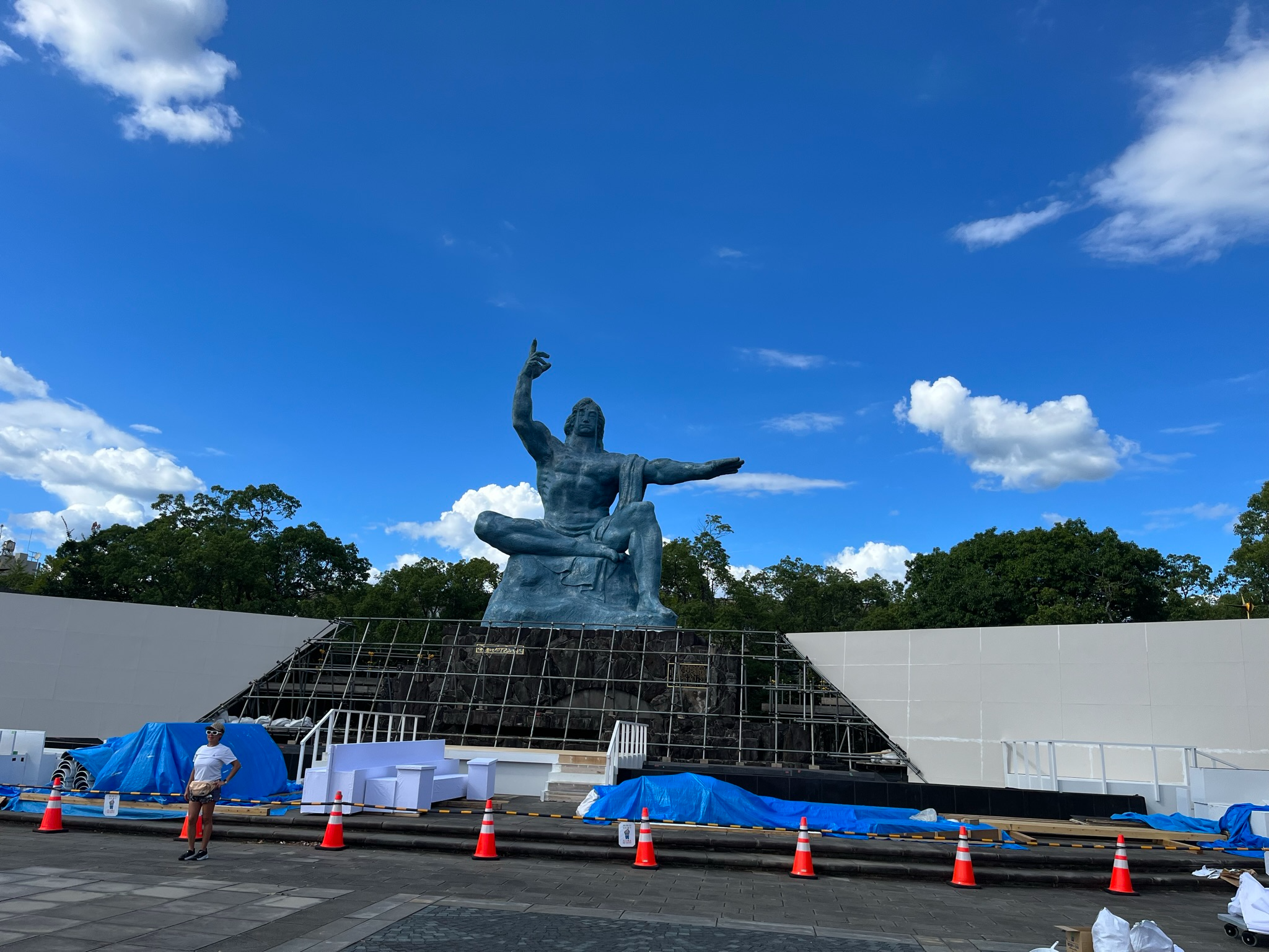 The Nagasaki Peace Statue in Peace Park, a large bronze figure seated on a stone pedestal with one arm raised and the other extended beneath a bright blue sky.